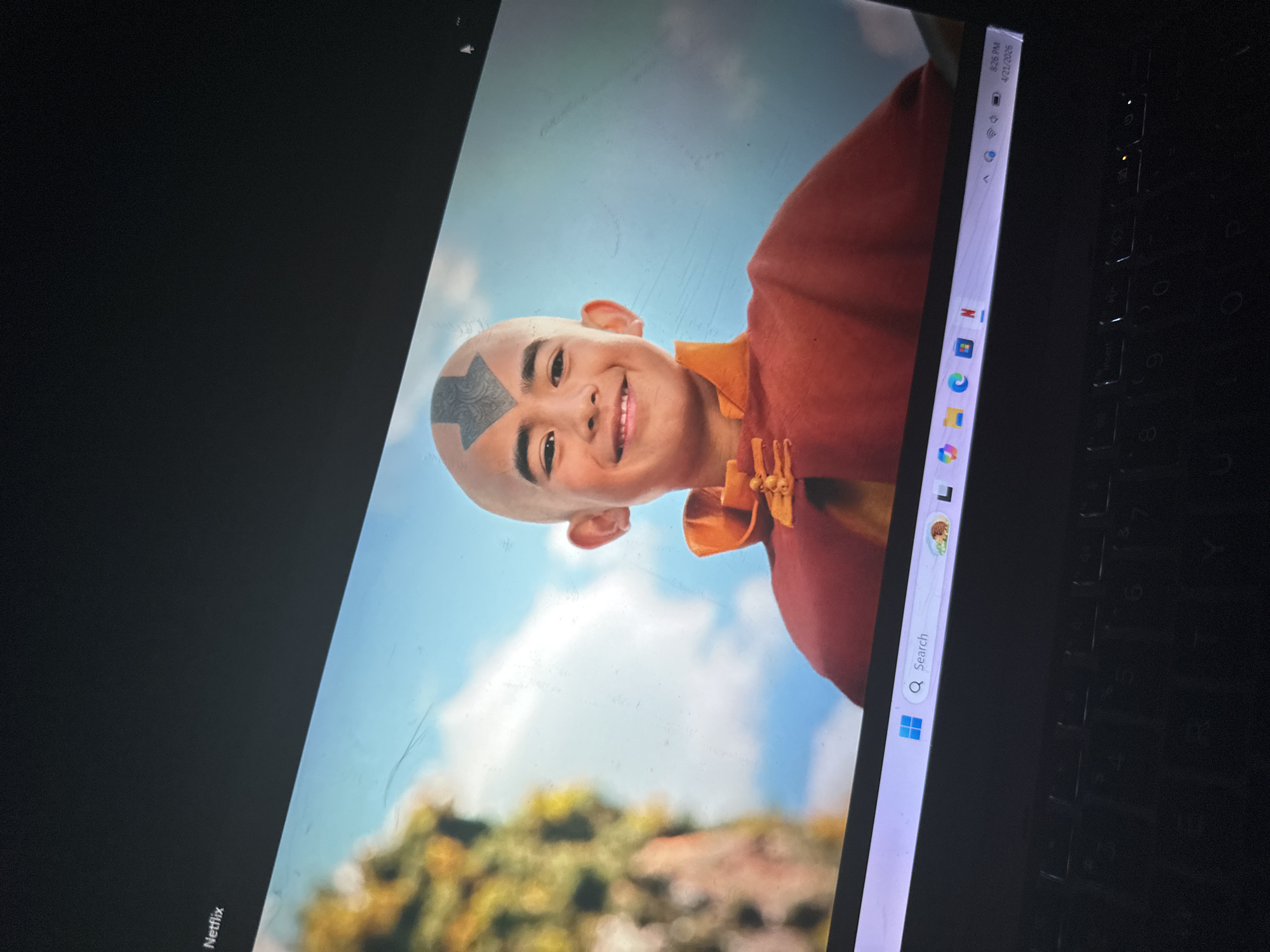 The image displays a laptop screen, rotated clockwise, showcasing a scene from a live-action adaptation of Avatar: The Last Airbender. The central subject is a young boy portraying Aang, characterized by his bald head and the distinctive blue arrow tattoo on his forehead. He wears a traditional orange and red outfit, consistent with Air Nomad attire, and is captured in a cheerful pose, smiling broadly with a happy expression while looking slightly upwards and off-screen.
The background visible on the screen is an outdoor setting under a clear, bright blue sky with soft white clouds. Blurred green foliage in the lower portion suggests a natural environment, possibly a hill or forest. The laptop's Windows taskbar is prominent along the right edge of the screen, displaying the time "8:26 PM" and the date "4/21/2025". Several application icons are visible, including those for Microsoft Edge, File Explorer, Mail, Photos, and Netflix. A search bar with the text "Search" is also present. The word "Netflix" is also visible, rotated, in the bottom left corner of the screen. A section of the laptop's black keyboard is partially visible to the right of the screen. The screen itself shows signs of use, with visible dust, smudges, and fine scratches. This image was captured in Kirana, Nigeria.