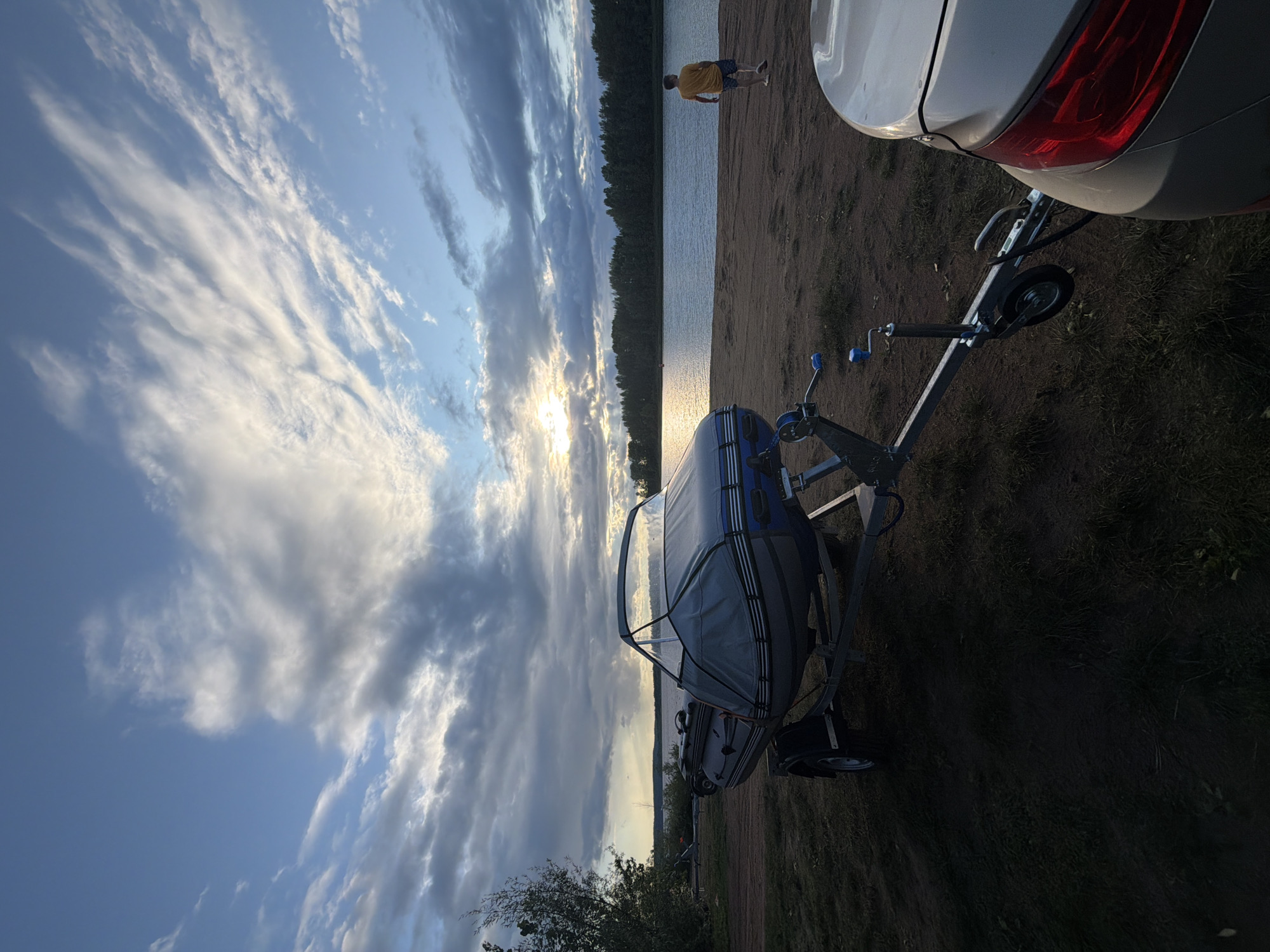 This image depicts a sunset scene by a body of water, likely the Kama River, near the city of Chaikovsky, Russia. A grey inflatable boat on a trailer is parked on a sandy shore in the foreground. A portion of a silver car is visible on the right, attached to the boat trailer. Further back, a person wearing a yellow shirt and shorts walks away from the camera towards the water. The sky is filled with dramatic clouds, with the sun partially visible through them, casting a warm glow on the water and the distant tree line. The overall impression is one of a serene evening by the river.