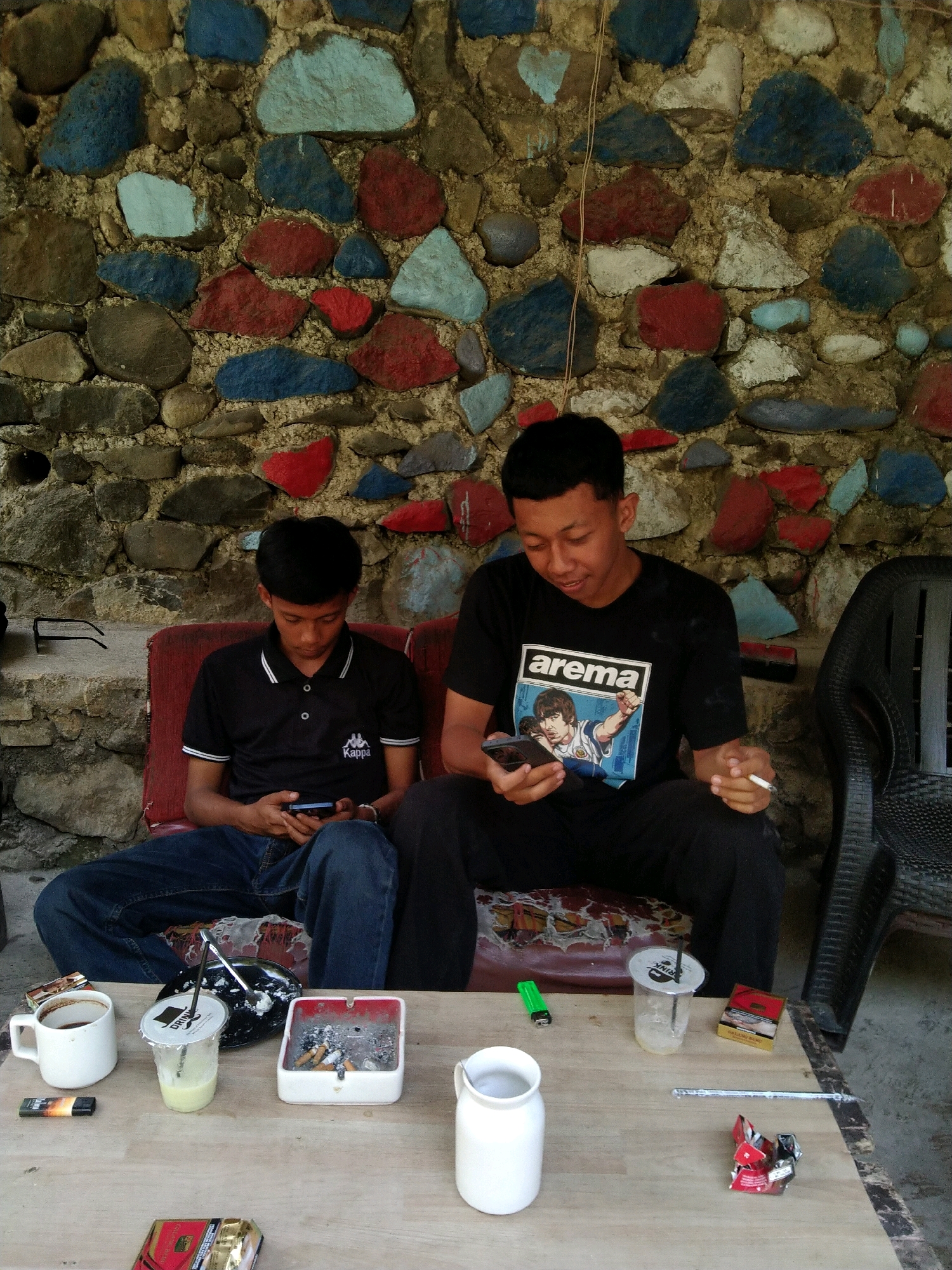 Two young Indonesian men are seated next to each other on a red cushioned bench. The man on the left is wearing a black polo shirt with white trim and blue jeans, and he is looking down at his mobile phone. The man on the right is wearing a black t-shirt with a graphic of two men on it, black pants, and is holding a cigarette in his right hand while looking at his mobile phone. Both men are positioned in front of a vibrantly colored stone wall, with stones painted in red, blue, and light blue. In front of them is a wooden table laden with various items. These include a white mug of coffee, a plastic cup with a straw and a milky-colored drink, a cigarette lighter, an ashtray filled with cigarette butts, a pack of cigarettes, and a white ceramic jar. The overall setting appears to be an outdoor seating area, possibly a cafe or roadside stall, in Malang, Indonesia.
