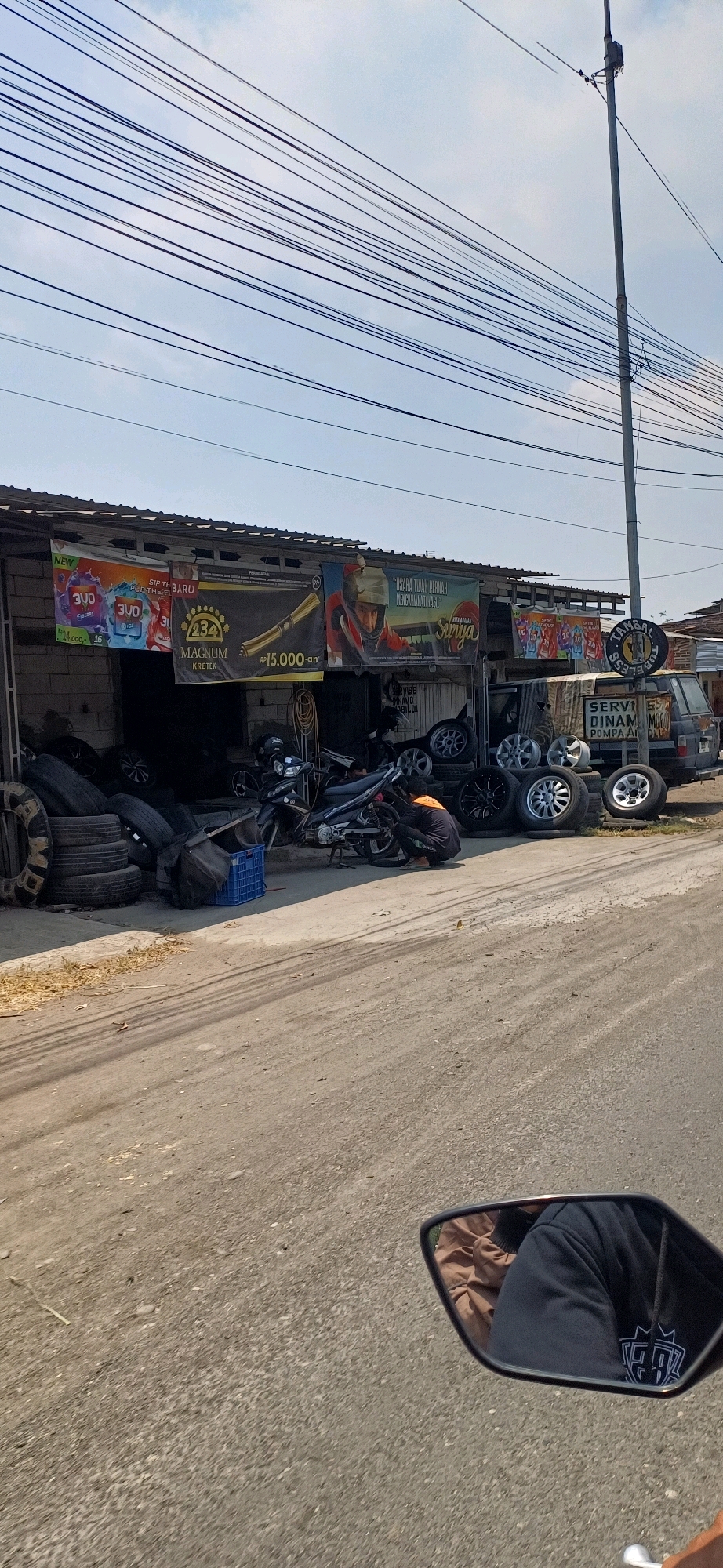 In the city of Kecamatan Pati, Indonesia, a storefront with an open-air covered area displays numerous tires and wheels, suggesting a tire shop or repair service. A black motorcycle is parked in front of the shop, and a person wearing a dark shirt and orange jacket is crouched down, appearing to work on the motorcycle's rear wheel. Banners with colorful graphics and text are hung across the front of the building. A dark-colored van is parked to the right of the shop. The ground in front of the shop is paved and shows tire marks. Overhead, a complex network of power lines stretches across a clear blue sky. In the foreground, a motorcycle's side mirror reflects the image of a person wearing a dark hoodie and brown jacket.