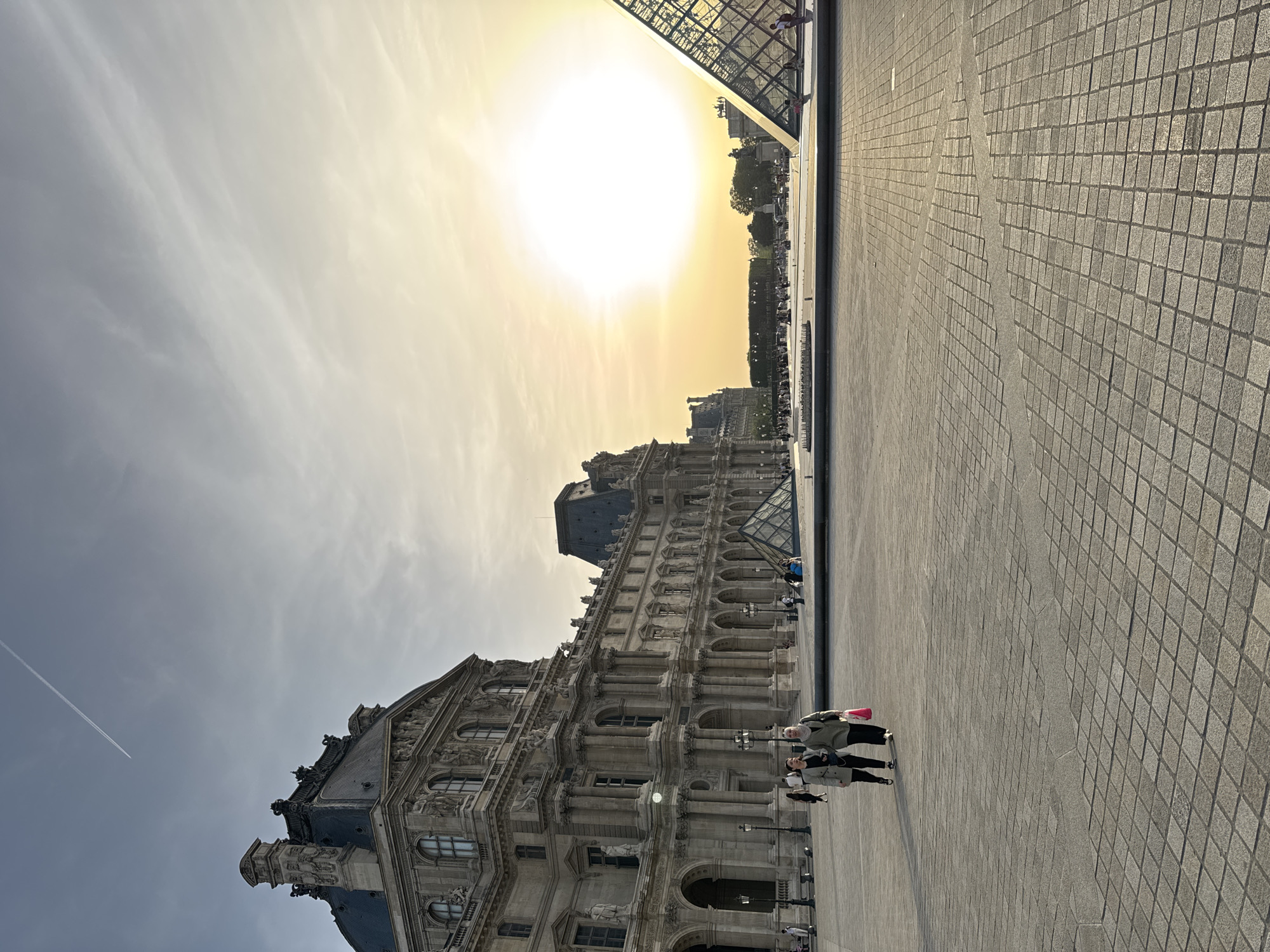 The image shows a view of the Louvre Museum in Paris, France. The foreground is a stone-paved courtyard. Two women are walking on the right of the frame. The Louvre Palace is visible behind them, with its ornate facade, arches, and classical sculptures. The Louvre Pyramid is partially visible at the top right, reflecting the setting sun. The sky is filled with clouds, with a bright sun casting a golden light over the scene. A contrail from an airplane cuts across the sky.