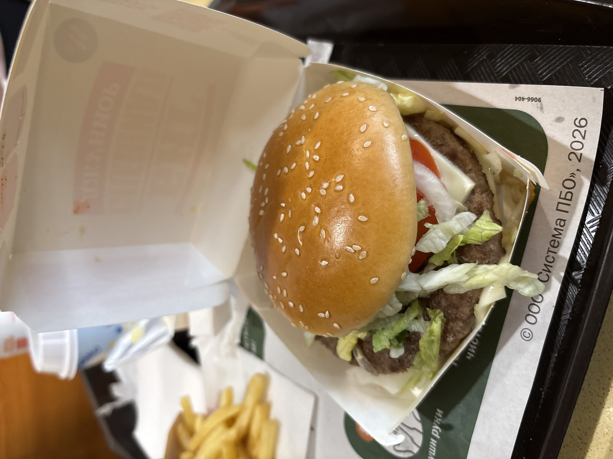 This media file presents an overhead view of a fast-food meal, likely consumed indoors at a restaurant or food court. The main subject is a hamburger, neatly arranged in an open cardboard box. The burger features a sesame seed bun, two beef patties, melted cheese, a slice of tomato, white onion, and shredded lettuce.
To the bottom left, a portion of golden french fries in a paper holder is partially visible, indicating a complete meal. All items are placed on a black, textured fast-food tray. Another white paper cup or container is faintly visible on the far left.
Notable details include Russian text visible on the paper packaging. On the right side of the burger box's exterior, the text "© ООО Система ПБО, 2026" is present, along with a number "9066-404" printed upside down. Faint, reversed text inside the burger box appears to be "БУРГЕР КИНГ" (Burger King), suggesting the meal is from that particular chain. A partial phrase "чистые руки" (clean hands) is also discernible on a wrapper or napkin beneath the burger box. The overall scene depicts a casual dining experience in Saint Petersburg, Russia.