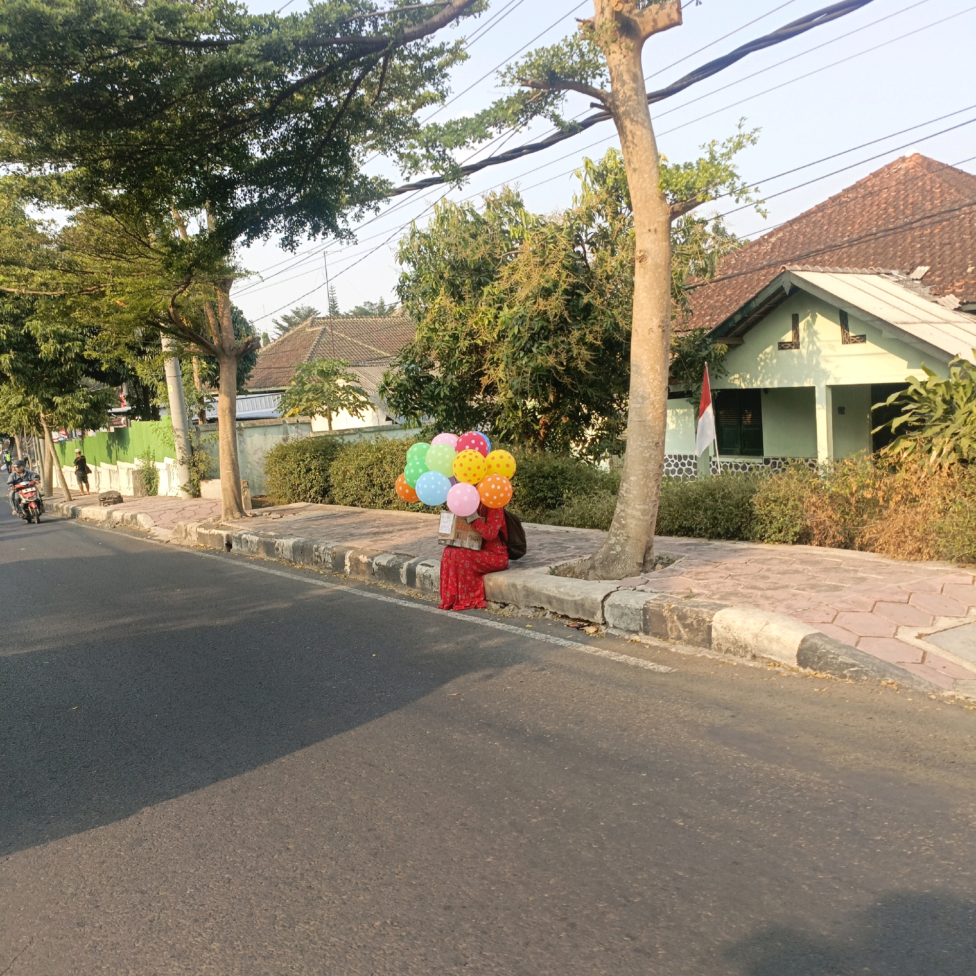 The image depicts a street scene in Salatiga, Indonesia, during daylight hours. In the foreground, a paved road stretches across the frame, with a sidewalk adjacent to it. The sidewalk is bordered by a curb and a stone wall. A person, wearing a red dress and a backpack, is seated on the curb, holding a bunch of colorful balloons. The balloons are round and varied in color, including red, orange, yellow, green, and blue, some with white polka dots. Behind the sidewalk, a light green house with a tiled roof is visible, featuring a porch and a red and white flag. Trees with lush green foliage are scattered throughout the scene, providing shade. Power lines crisscross the sky above. A motorbike is seen on the road in the background, with two individuals on it. The overall atmosphere suggests a quiet, residential street in a tropical climate.