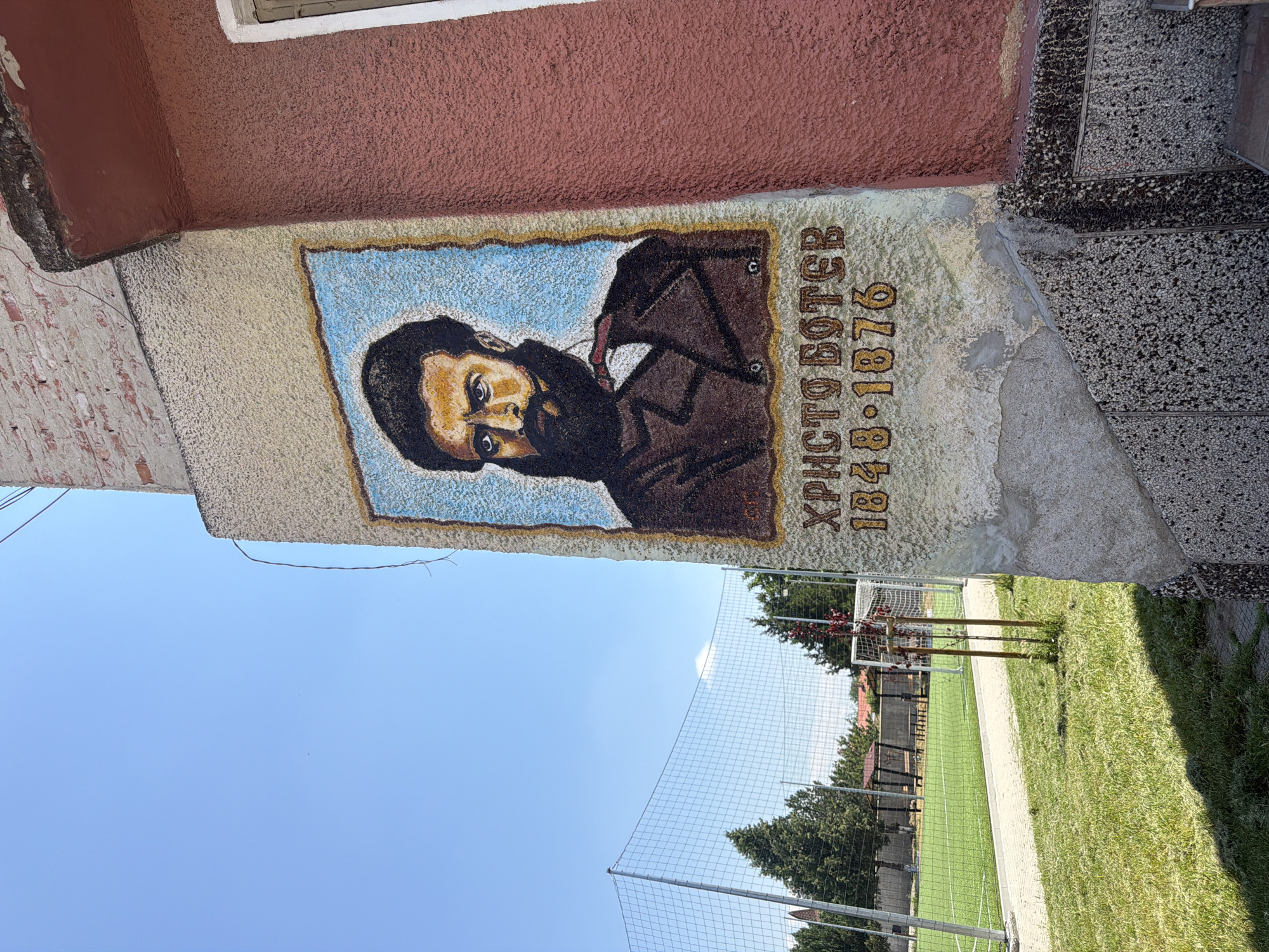 The image shows a mural painted on the side of a building in Zlatitrap, Bulgaria. The mural depicts a portrait of a man with a beard and dark hair, wearing a dark suit and a white shirt with a red tie. Below the portrait, the name "ХРИСТО БОТЕВ" and the years "1848-1876" are painted in yellow. The building appears to be made of brick and stucco, with a red-tiled roof visible at the top. To the left of the building, there is a grassy area with a fence and a soccer field in the background. The sky is clear and blue.