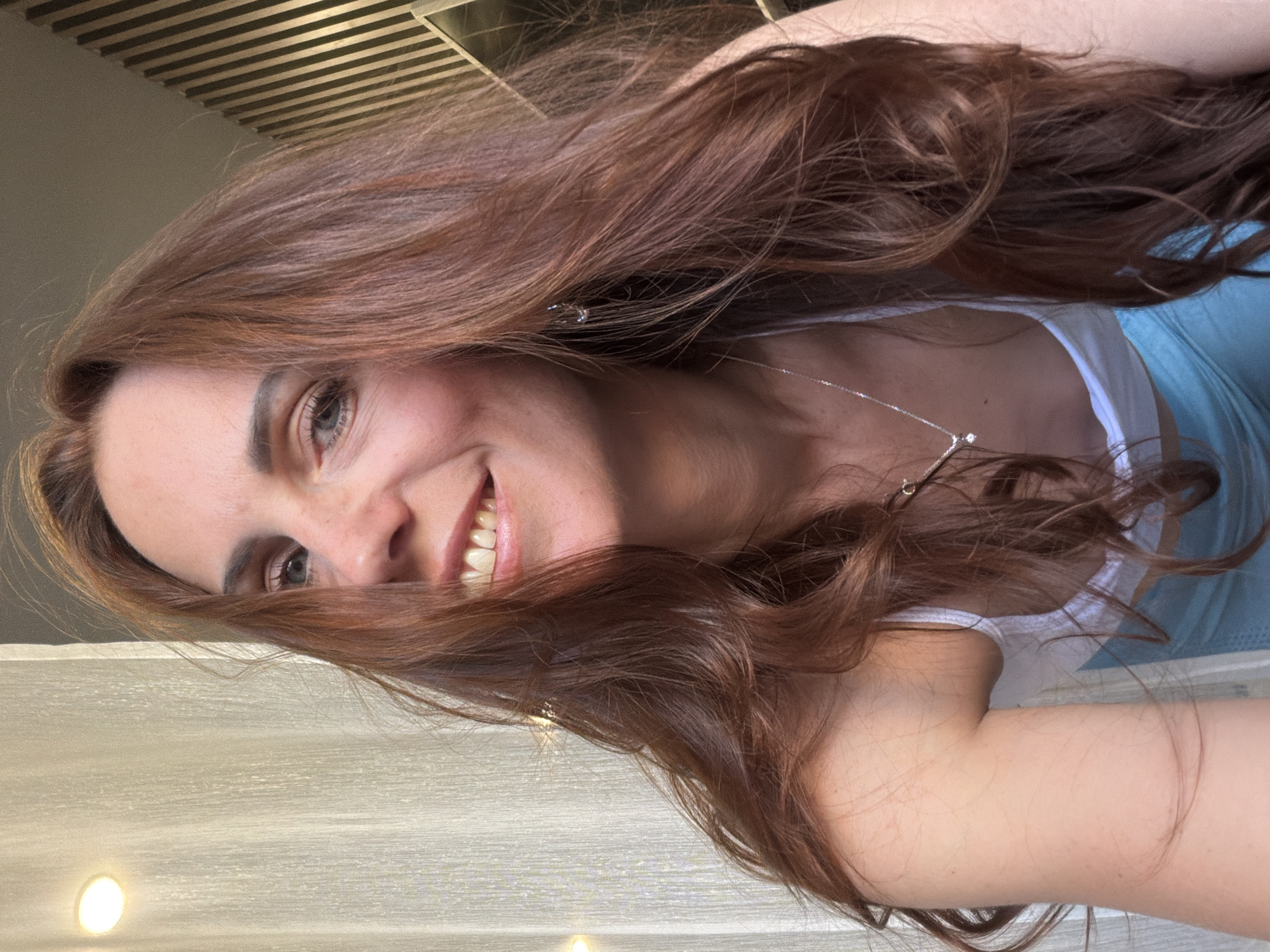 A close-up portrait shows a woman with long, wavy, reddish-brown hair, smiling broadly at the camera. She has fair skin and light-colored eyes, with her teeth visible in her joyful expression. She is wearing a white top or camisole beneath a light blue garment, possibly a tank top or swimwear. A thin silver chain necklace with a small, clear pendant hangs around her neck, partially obscured by her hair. A delicate silver earring is visible in her left ear.
The image is taken from a slightly low angle, focusing primarily on her face and upper body. The background features a slatted ceiling structure above her, suggesting an indoor setting. Below her, there is a light-colored, possibly textured wall or surface with at least two circular light fixtures. The lighting is bright and appears even. There is no visible text in the image. The image file itself is oriented 90 degrees counter-clockwise.