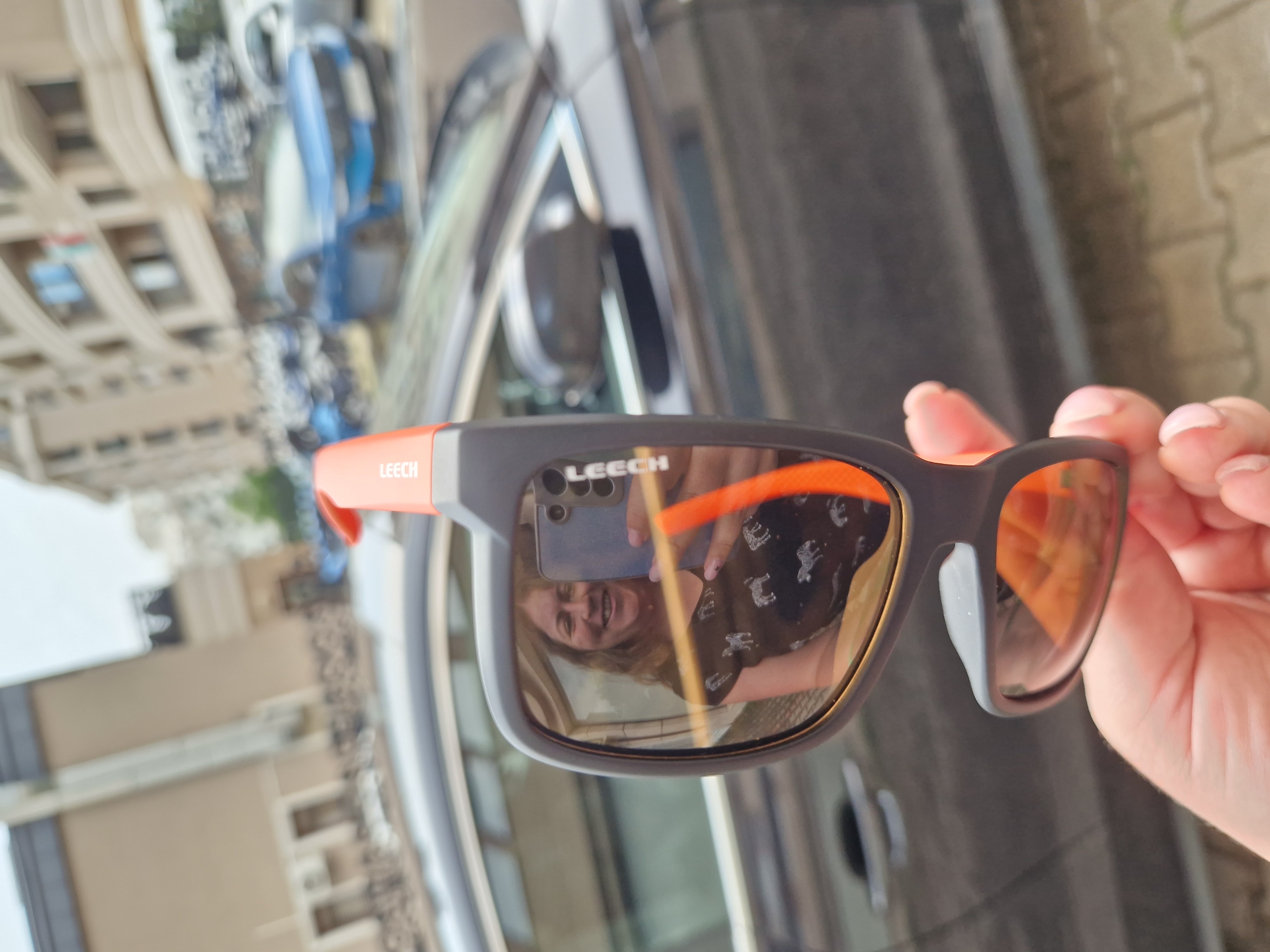 This is a close-up, vertically oriented photo of a person's hand holding a pair of sunglasses. The sunglasses have dark gray frames with bright orange accents and orange tinted lenses. The left lens reflects a scene that includes a building with multiple floors and balconies, suggesting an urban setting. A blue vehicle is also visible in the reflection. The right lens reflects the person holding the sunglasses, who appears to be a woman with blonde hair, smiling. She is wearing a dark t-shirt with a pattern of white animals. The "LEECH" logo is visible on both the frame of the sunglasses and in the reflection. The lighting suggests it is daytime, likely sunny, given the reflection and bright orange lenses. The paving stones visible in the background of the main image indicate an outdoor setting. Based on the provided location context, the scene is likely in Sofia, Bulgaria.