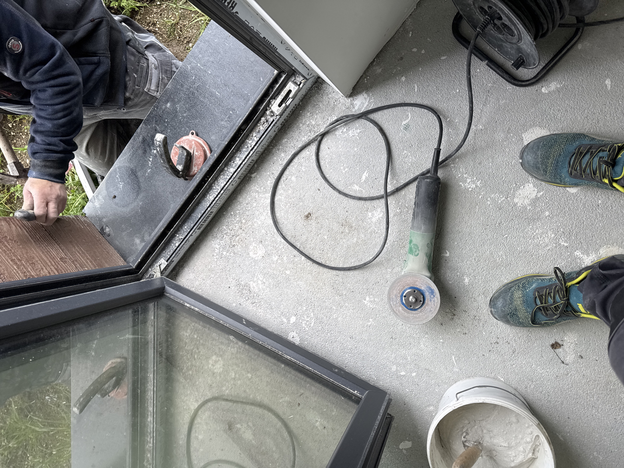 The image shows a construction or renovation scene. A person is visible on the left, wearing a dark jacket and grey pants. They appear to be installing or adjusting a dark-framed glass panel or window. To the right of the glass panel is a grey angle grinder with a grinding wheel, lying on a grey concrete surface. Near the grinder, there's a bucket filled with a grey-ish substance, possibly plaster or cement. The feet of another person, wearing blue and yellow running shoes, are seen in the lower right of the image. A black coiled extension cord lies near the upper right corner. The location cannot be confirmed based on the image alone, but it is requested to be in Burgas, Bulgaria.