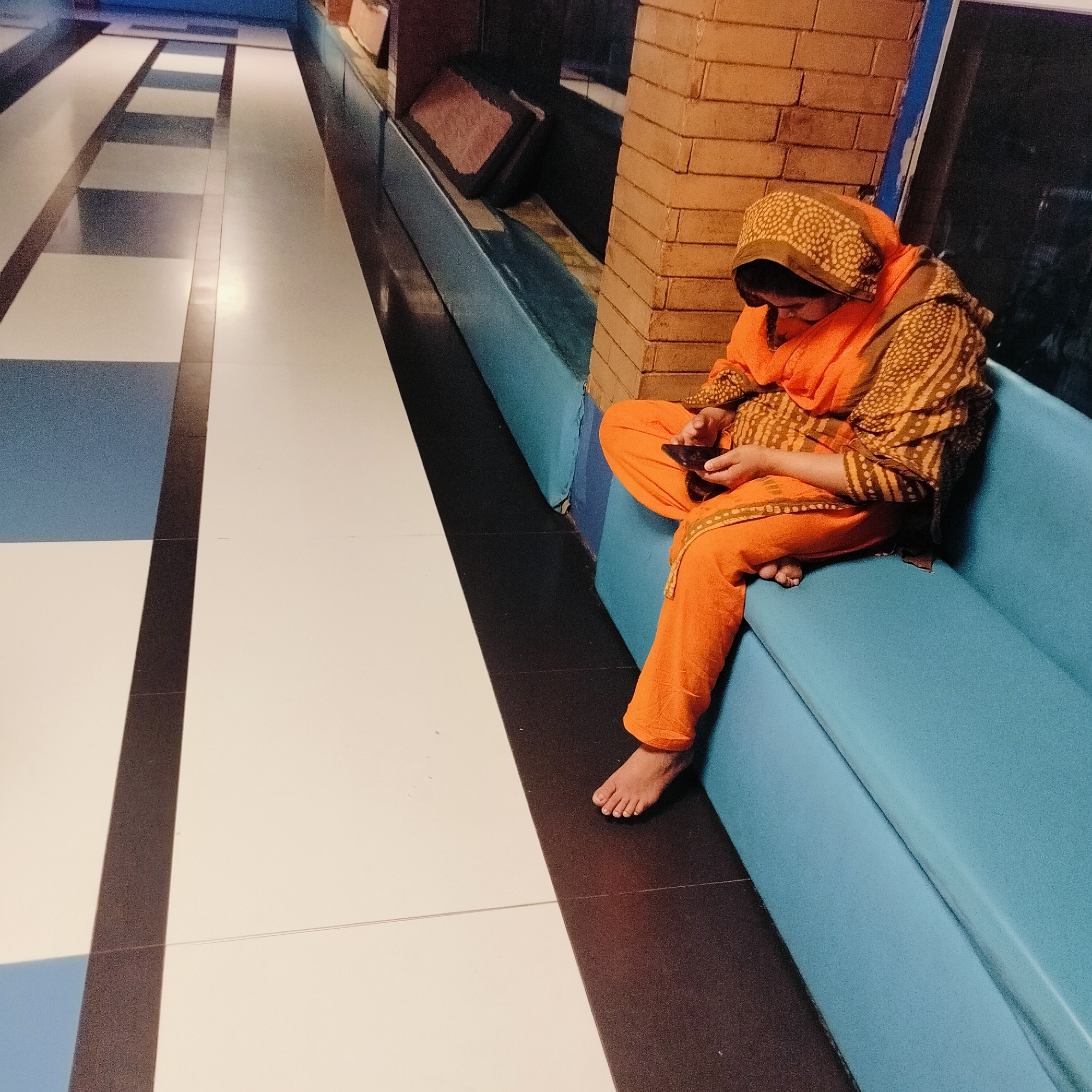 A woman sits on a light blue cushioned bench in what appears to be a public waiting area. She is dressed in an orange patterned salwar kameez with a matching head covering. She is barefoot and looking down at a mobile phone in her hands.
The bench is positioned against a wall partially covered with orange brick-like tiles. The floor is tiled in a linear pattern of alternating white, blue, and dark gray rectangles. The perspective suggests a long hallway or waiting room.
Given the attire and context, it is reasonable to infer that this image was taken in a South Asian location, and the prompt specifies the city of Dhaka, Bangladesh.