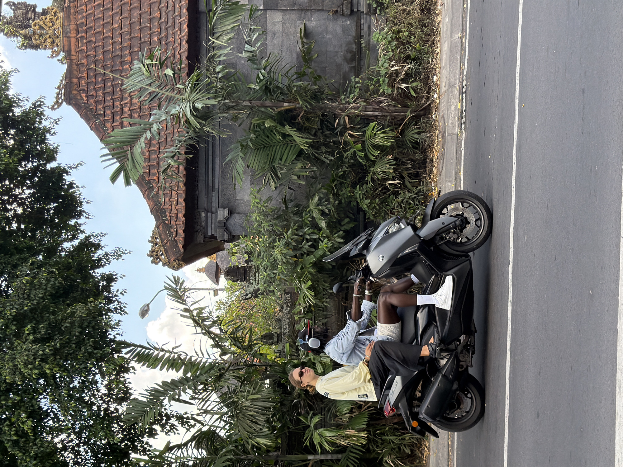This image depicts an outdoor scene in North Kuta, Indonesia, featuring two individuals on a dark grey Yamaha XMAX scooter on the side of a paved road. Lush tropical vegetation, including palm fronds and dense trees, frames the left side of the scene.
The person driving the scooter is a dark-skinned man wearing a light blue denim-style shirt with "NEMIS" printed on it, white shorts, white socks, and white sneakers. He has dark hair, possibly dreadlocks, and wears black headphones and a black baseball cap, looking straight ahead. Several bracelets adorn his left arm.
Behind him, the passenger, a light-skinned individual, wears a light yellow long-sleeved shirt, dark pants, and black shoes. They have light hair and are wearing sunglasses, looking slightly to their left.
In the background, traditional Indonesian architecture is visible, characterized by a red tiled roof with ornate golden carvings and dark grey stone walls, set amidst more tropical foliage. A street light is discernible against a partly cloudy blue sky. The bright, clear daylight suggests the time of day is morning to early afternoon. The overall atmosphere appears relaxed.