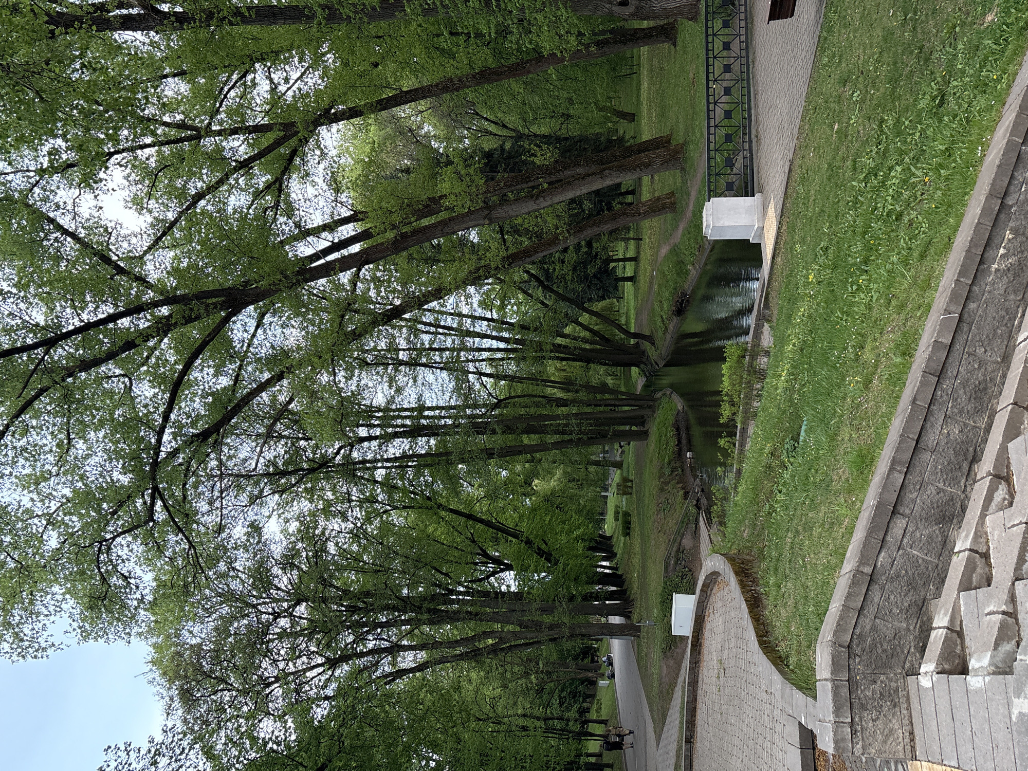 The image depicts a park scene in Minsk, Belarus. A canal runs through the center, lined with trees on both sides, their branches forming a canopy overhead. On the right side of the canal is a grassy bank, bordered by a paved walkway. A stone wall and steps are visible in the foreground on the right. Along the walkway are benches. Several people can be seen walking along the paved path. The image is taken during the daytime with natural lighting.