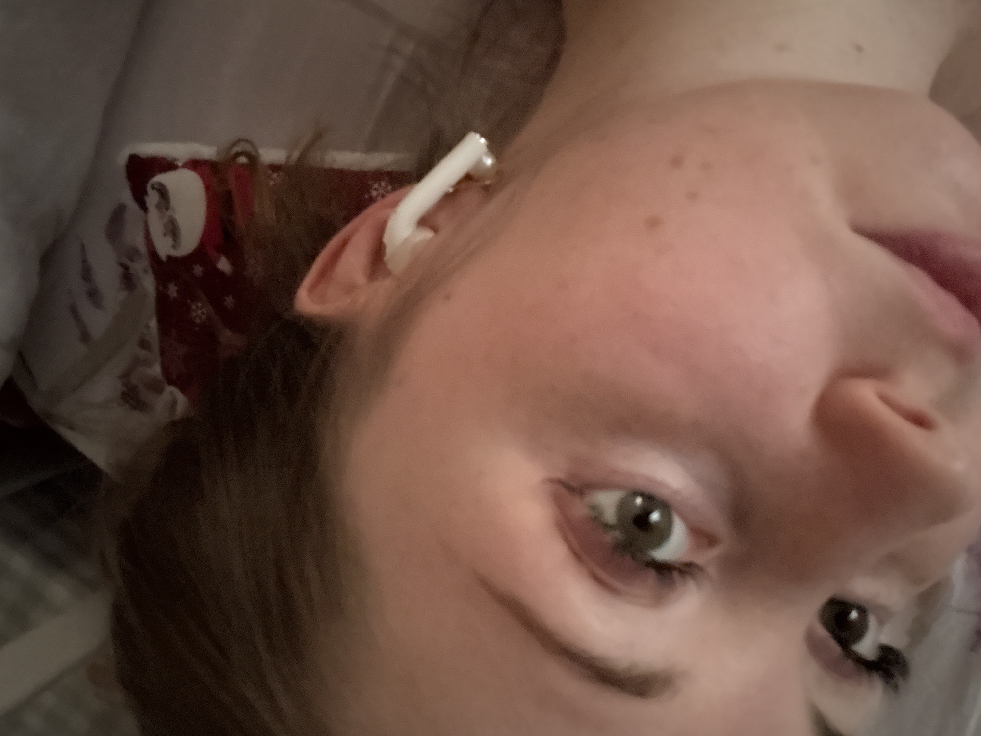 A close-up, slightly angled shot of a person's face, presumably female. The person has fair skin, freckles, and light brown hair. They have green eyes and are wearing what appears to be an earbud in their left ear. The background is blurred, but there's a glimpse of patterned fabric, possibly a blanket or pillow. The location is unknown.