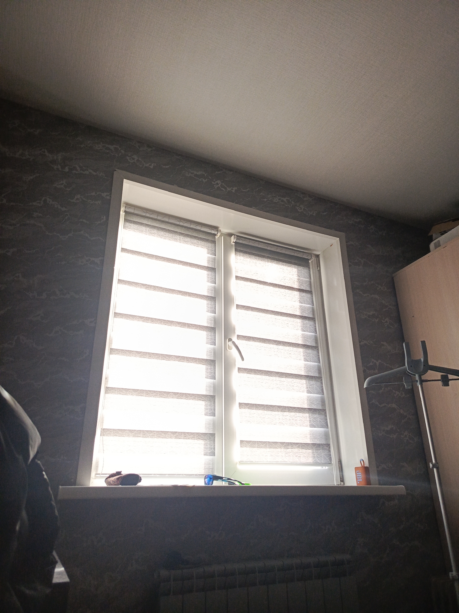 The image captures an interior scene, likely a room in an apartment, with a window as the central focus. The window is equipped with dual roller blinds, one transparent and one opaque, creating a striped effect as light passes through. The blinds are a modern design, alternating between sheer and solid fabric. A white windowsill beneath the window holds a few items: a dark-colored object, possibly a pair of sunglasses with blue lenses, and a small orange rectangular object with writing on it, possibly a timer or a small container.
The walls are covered in dark wallpaper with a patterned texture, featuring an abstract, vein-like design in a lighter grey. The ceiling is painted a light, neutral color. To the right of the window, a portion of a wooden closet or wardrobe is visible, along with a metallic clothes rack. The overall lighting suggests it is daytime, with sunlight filtering through the blinds, creating a bright and somewhat hazy atmosphere. The lower part of the image shows a section of a radiator, suggesting the presence of a heating system.
Without any external clues within the image, it is impossible to definitively pinpoint the location to Lesosibirsk, Russia. However, assuming this context is provided externally, the scene depicts a common domestic setting that could be found in many residential areas.