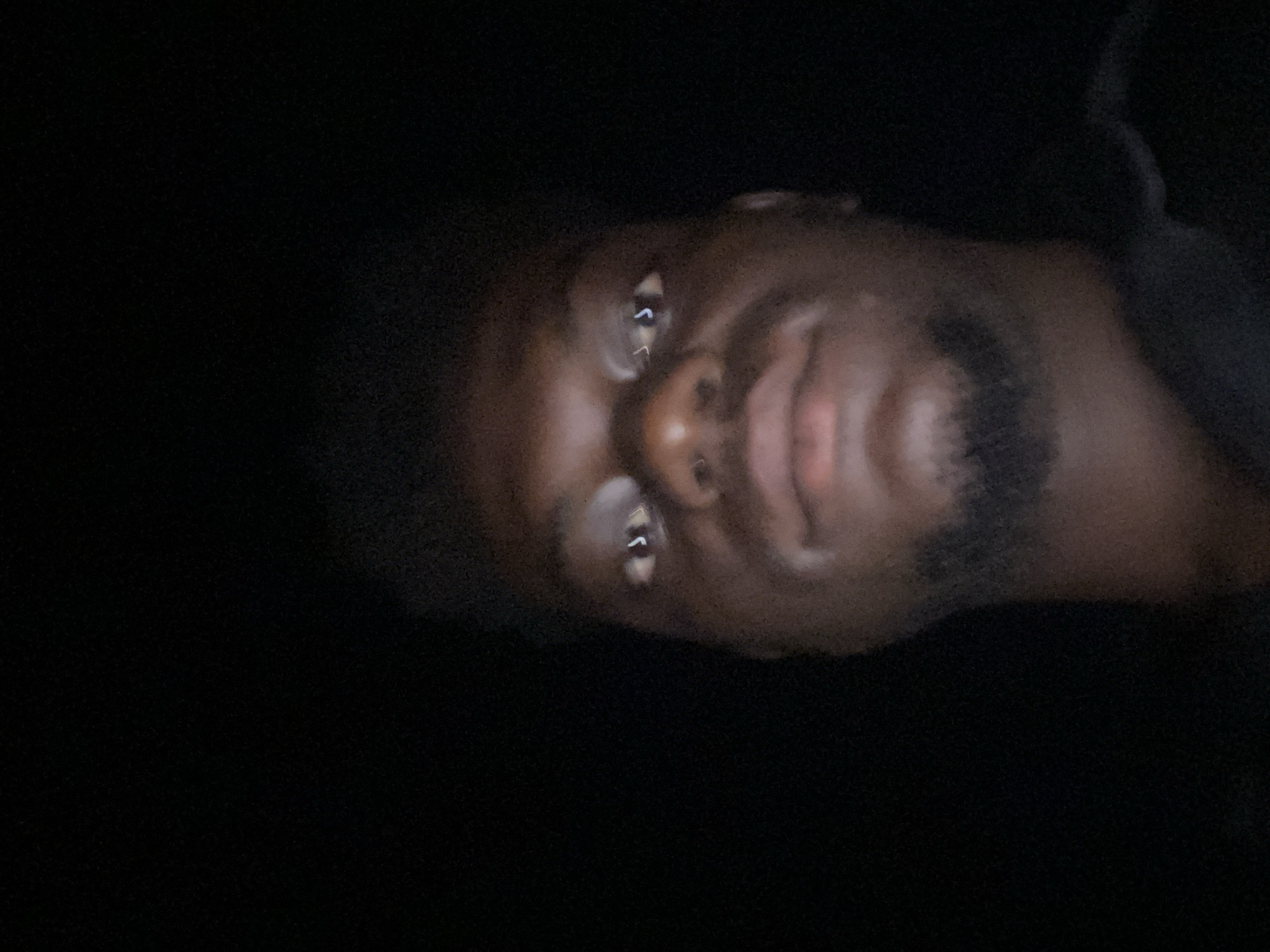 A close-up, low-light image features a person's face, likely a man, with dark skin, a beard, eyes reflecting light, and visible nose and lips. The background is mostly black, suggesting a dimly lit environment. No specific city or location is discernible.