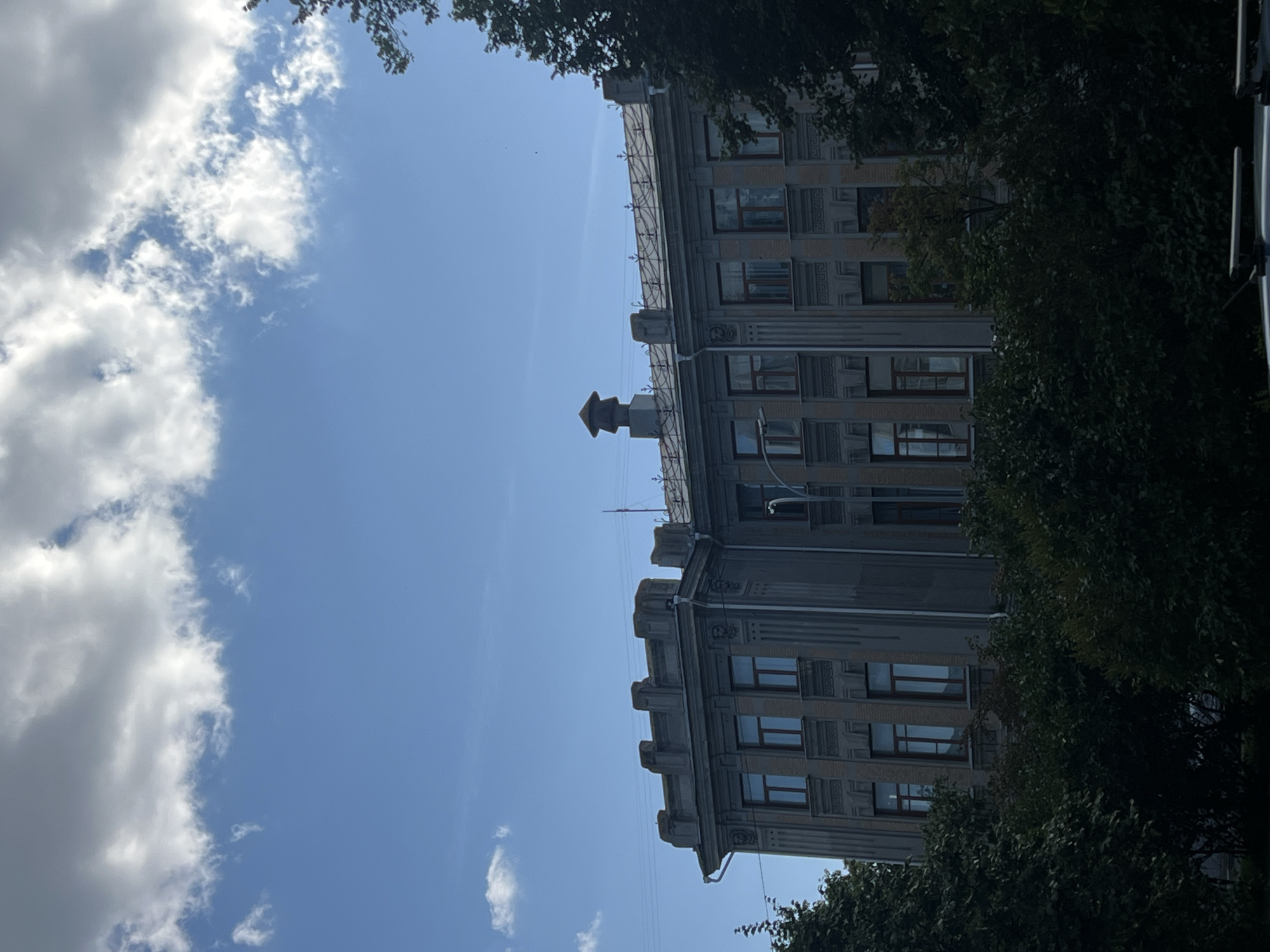 The image captures an overcast day with a bright blue sky visible above a light gray, multi-story building. The building features numerous rectangular windows with dark frames and a decorative cornice along the roofline. A ventilation shaft with a decorative cap sits on the roof. Dense green foliage in the foreground obscures the lower portion of the building. The scene appears to be an urban environment, possibly a street in Ulyanovsk, Russia, given the context provided.