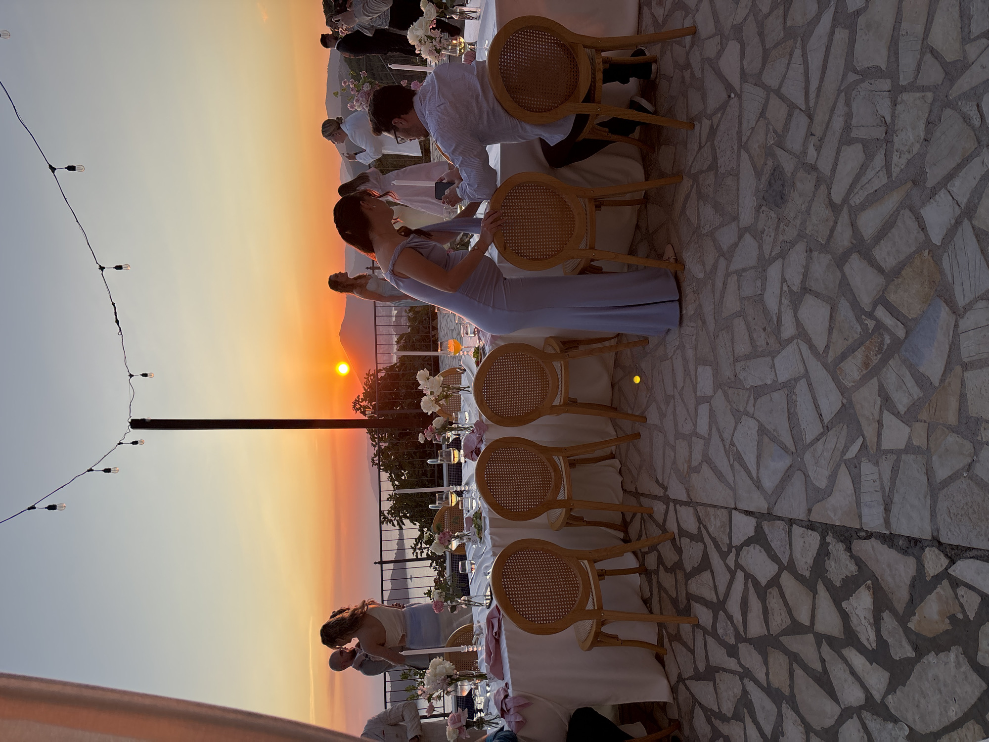 This image depicts an outdoor wedding reception at dusk, with a sunset visible in the background. The setting appears to be a terrace overlooking a body of water and hills. A long, white-draped table is set for a meal, with chairs arranged around it. String lights are hung overhead. Several guests are present, dressed in formal attire. A couple in the foreground is interacting, with one person holding a phone. The ground is paved with irregular stone tiles. The overall atmosphere suggests a celebration taking place during golden hour.