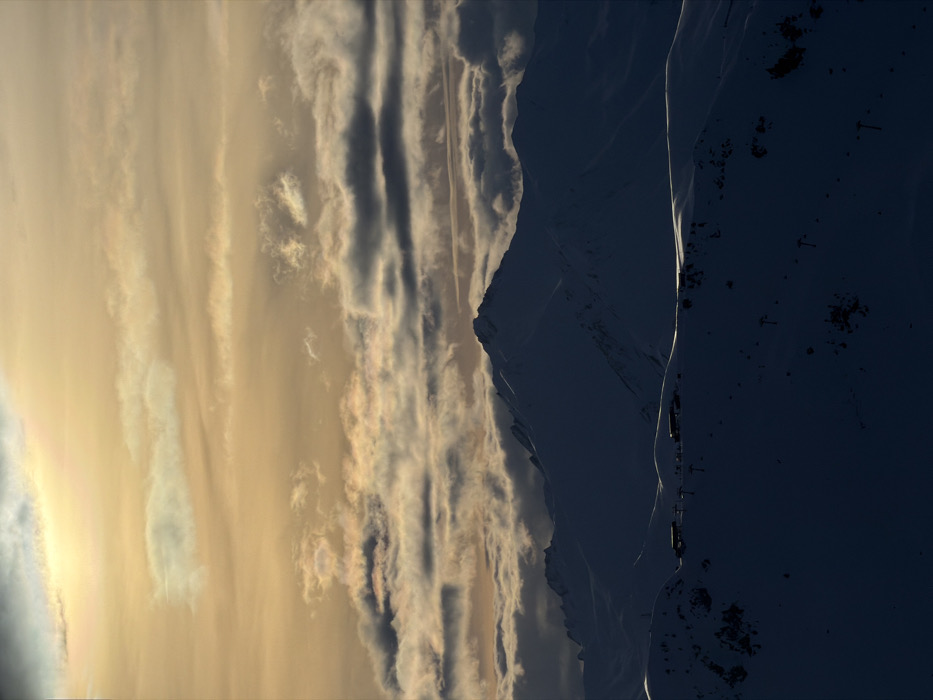 The image depicts a mountain range under a vibrant sky. The left side of the frame is filled with streaky clouds, colored in shades of golden yellow and white. The mountain range on the right is cast in deep shadows, revealing snowy peaks and sharp ridges. A vertical line of light highlights the boundary between the illuminated and shaded areas. Ski lifts are visible on the dark mountain side.