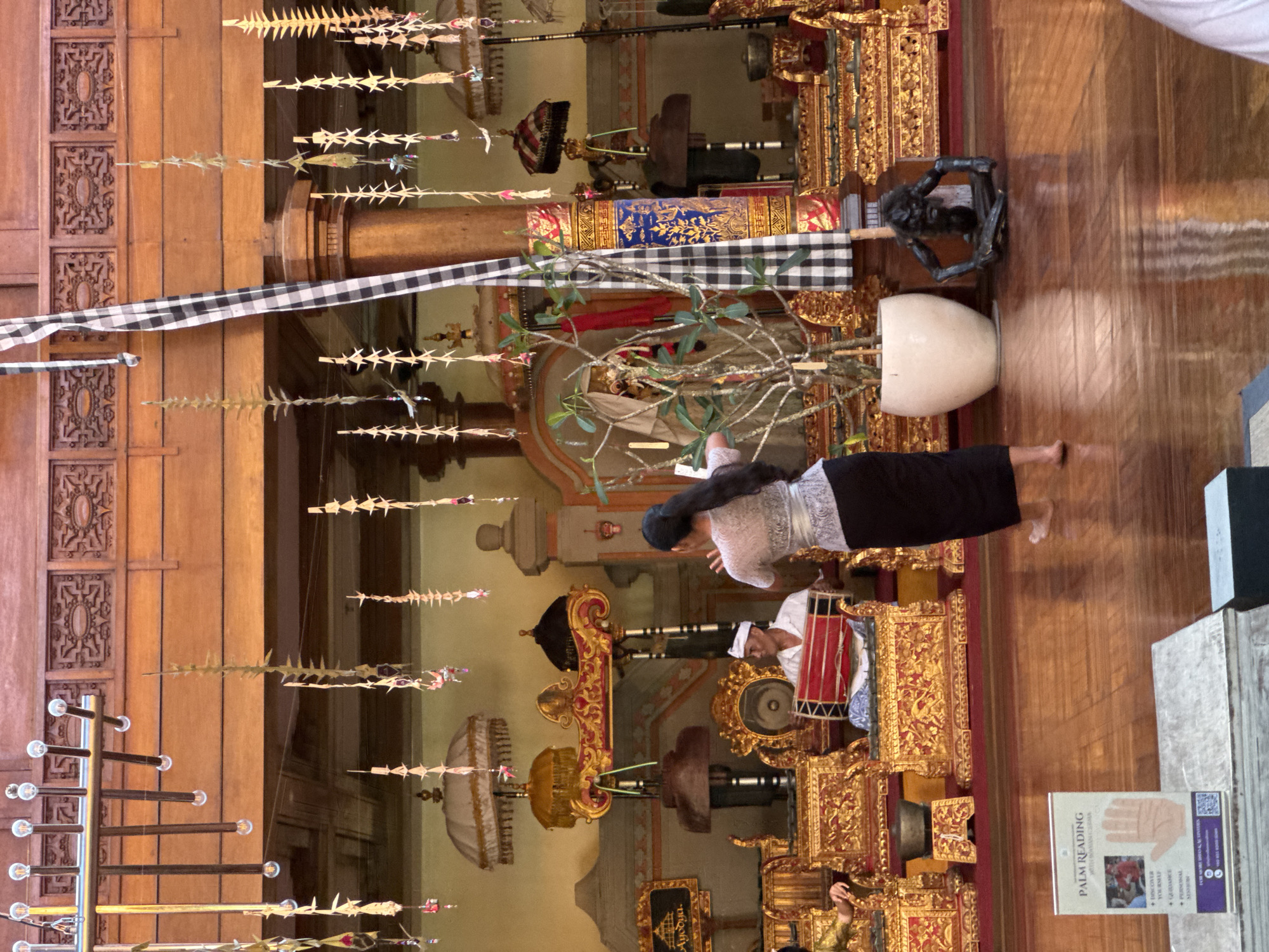 The image depicts an indoor cultural performance within an elaborately decorated space in Kuta Selatan, Indonesia. In the mid-ground, a female dancer, viewed from the back, with long dark hair tied back, wears a white lace-patterned top and a dark sarong, performing barefoot with her right arm raised. To her left, a male musician, wearing a white shirt and a white traditional headwrap (udeng), sits behind and plays a red barrel-shaped drum (kendang).
The background features multiple ornate, gold-colored tiered structures with red and black accents, serving as stands for various gamelan instruments, including gongs and bowl-shaped percussion. Several large white ceremonial parasols with gold fringes, along with smaller dark parasols, are positioned around these structures. A substantial wooden column on the right is partially wrapped in a black and white checkered fabric (saput poleng) and a richly patterned blue and gold textile. Above, numerous delicate, light-colored hanging decorations, possibly crafted from palm leaves, are suspended from a wooden ceiling adorned with carved panels. A modern chandelier with multiple exposed bulbs hangs in the upper left. In the mid-ground right, a potted plant with sparse branches sits in a white cylindrical planter, adjacent to a dark, squatting, human-like statue on the polished wooden floor. In the lower left foreground, a vertical white sign advertising "PALM READING" is visible, featuring a graphic of an open hand and textual details about services offered. The wooden floor is highly reflective.