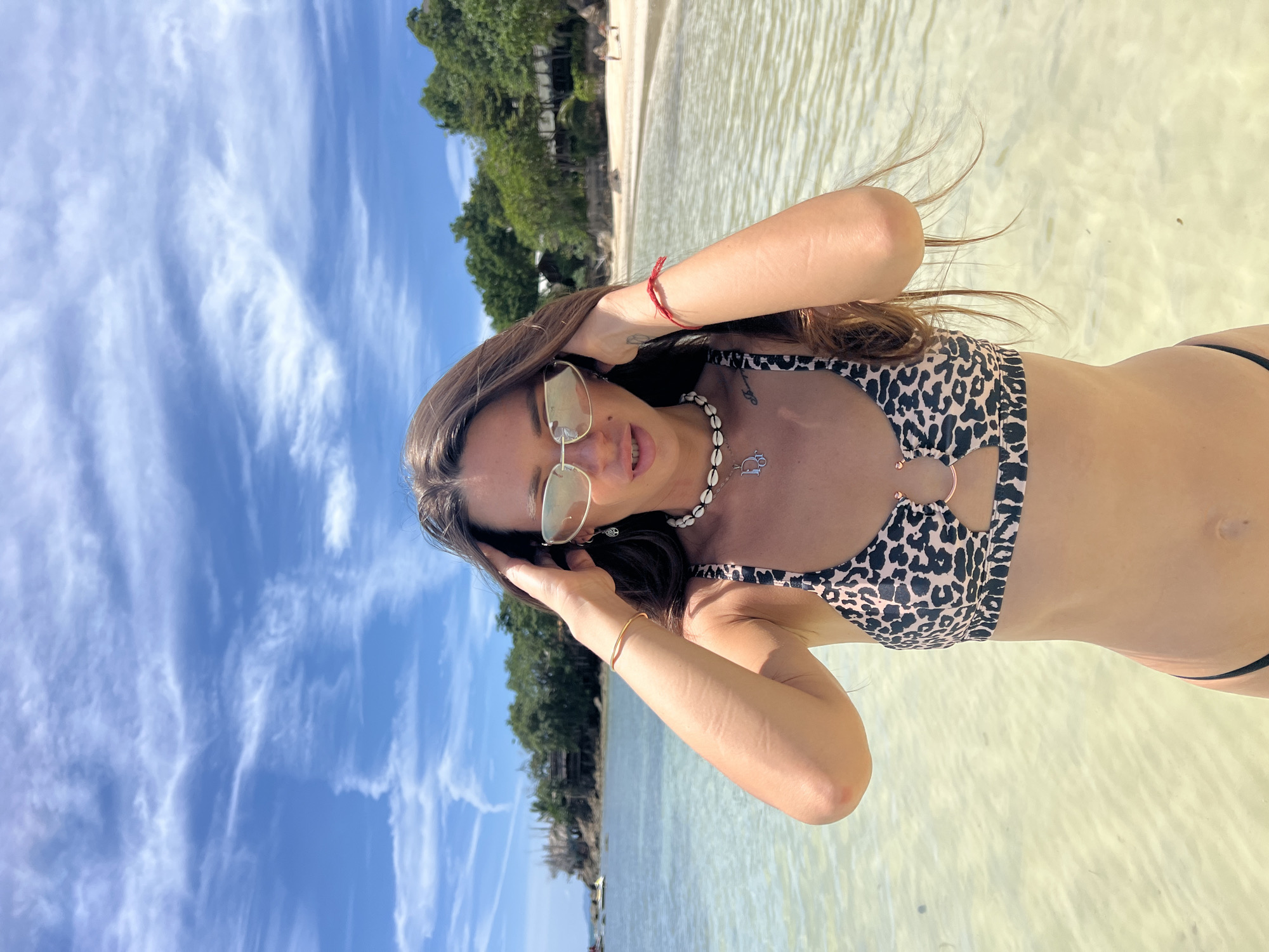 A woman stands in shallow, clear water on a beach. She is wearing a leopard print bikini and sunglasses. Her hands are raised to her hair, and she is looking at the camera. The background shows a sandy beach with trees and buildings. The sky is blue with scattered clouds. The scene is likely located on Koh Phangan, Thailand, given the prompt.
