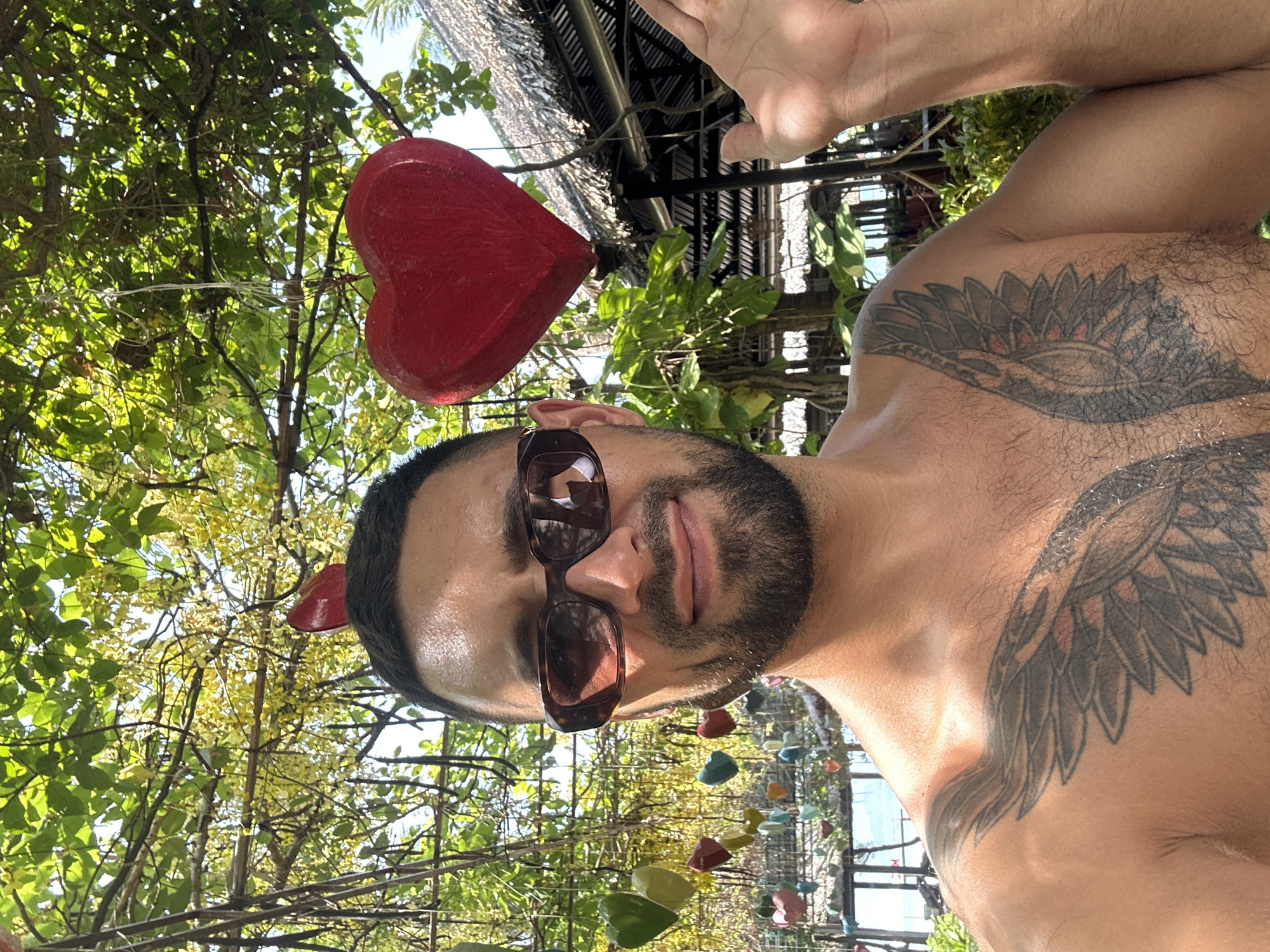 A man with a beard and sunglasses is posing for a selfie outdoors in Ninh Ích, Vietnam. He is shirtless and has a large tattoo of wings on his chest. He is standing under a tree with yellow flowers and decorative heart-shaped ornaments hanging from the branches. In the background, there is a thatched-roof building and the ocean. The man is smiling and has his left hand raised.
