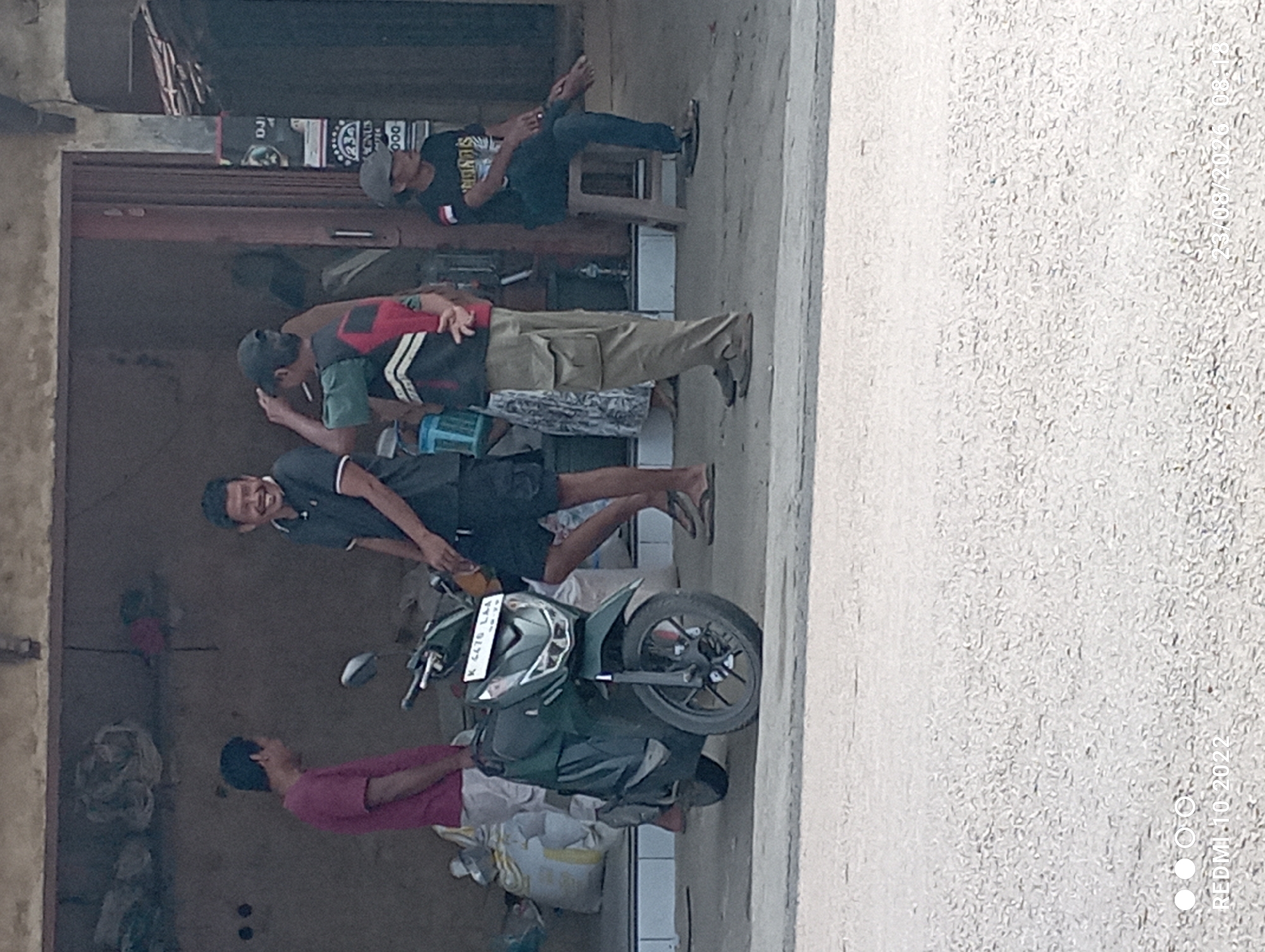 In Dukuhseti, Indonesia, several individuals are gathered outside a storefront. A dark green scooter is parked in the foreground. To the left, a man in a maroon t-shirt stands near a pile of sacks. A man in a dark grey polo shirt and black shorts is seen in the center, laughing. Another man, wearing a vest over a green t-shirt and khaki pants, stands with his hands behind his back. Further to the right, a man in a black t-shirt and blue jeans is seated on a stool, looking towards the right. The storefront appears to have a large open doorway. The scene is outdoors, with a concrete ground surface in front of the building.