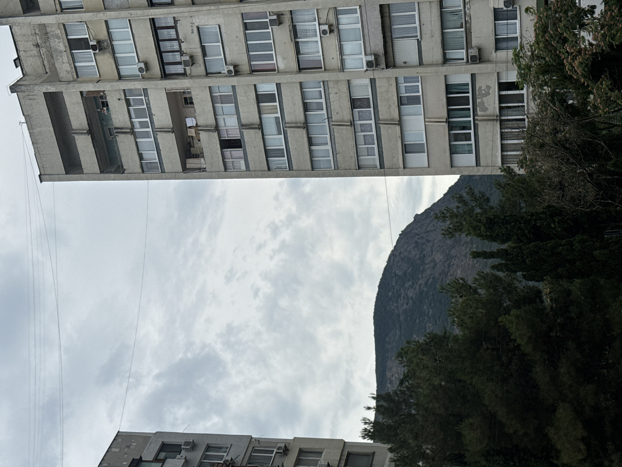 The image depicts a view from a balcony in the city of Partenit, Ukraine, looking towards a mountain and two apartment buildings. The apartment building in the foreground, on the right, is a multi-story concrete structure with numerous windows and balconies. Some of the windows are open, and air conditioning units are visible mounted on the exterior walls. The building on the left is partially visible and appears to be of similar construction. In the mid-ground, a lush green forest can be seen, and behind it, a rugged mountain rises against a cloudy sky. The overall scene suggests an urban environment surrounded by natural landscapes.
