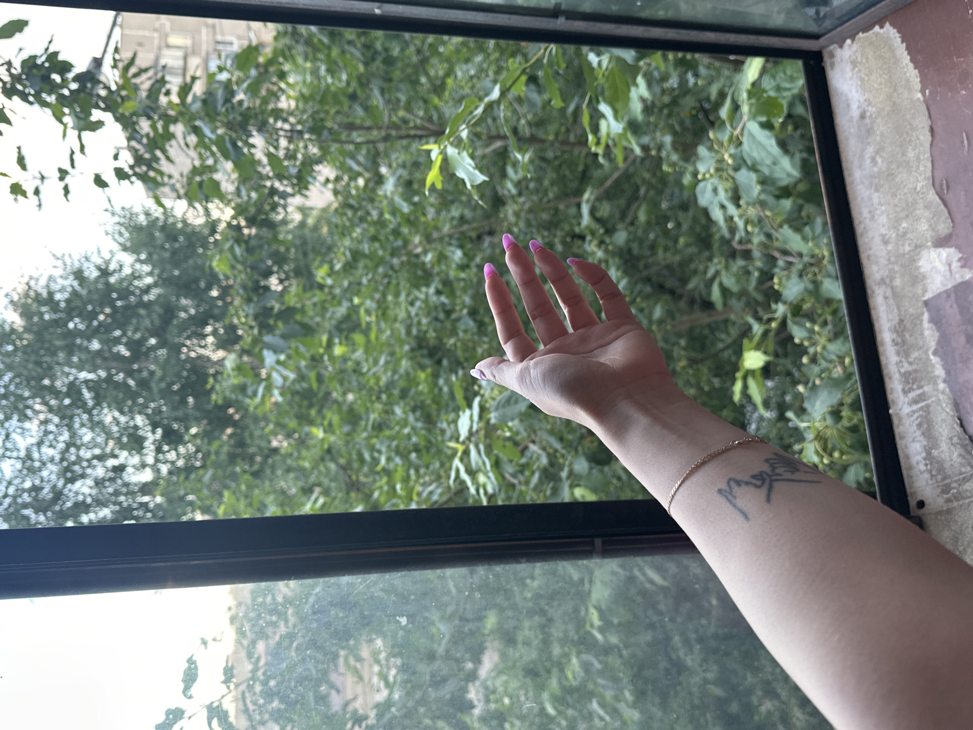 A person's arm and hand extend from the left side of the frame, reaching towards a window. The hand, with bright pink fingernails, is open and facing the viewer. A thin gold bracelet is visible on the wrist, and a black tattoo is present on the forearm. The scene appears to be an interior space looking out a window at a dense canopy of green trees and foliage. The window frame is dark and appears to be made of metal. Peeling paint or wallpaper is visible on the windowsill in the foreground. The image is captured from a medium-close shot, focusing on the arm and hand against the natural backdrop. The lighting suggests it is daytime. The location is specified as Magnitogorsk, Russia.