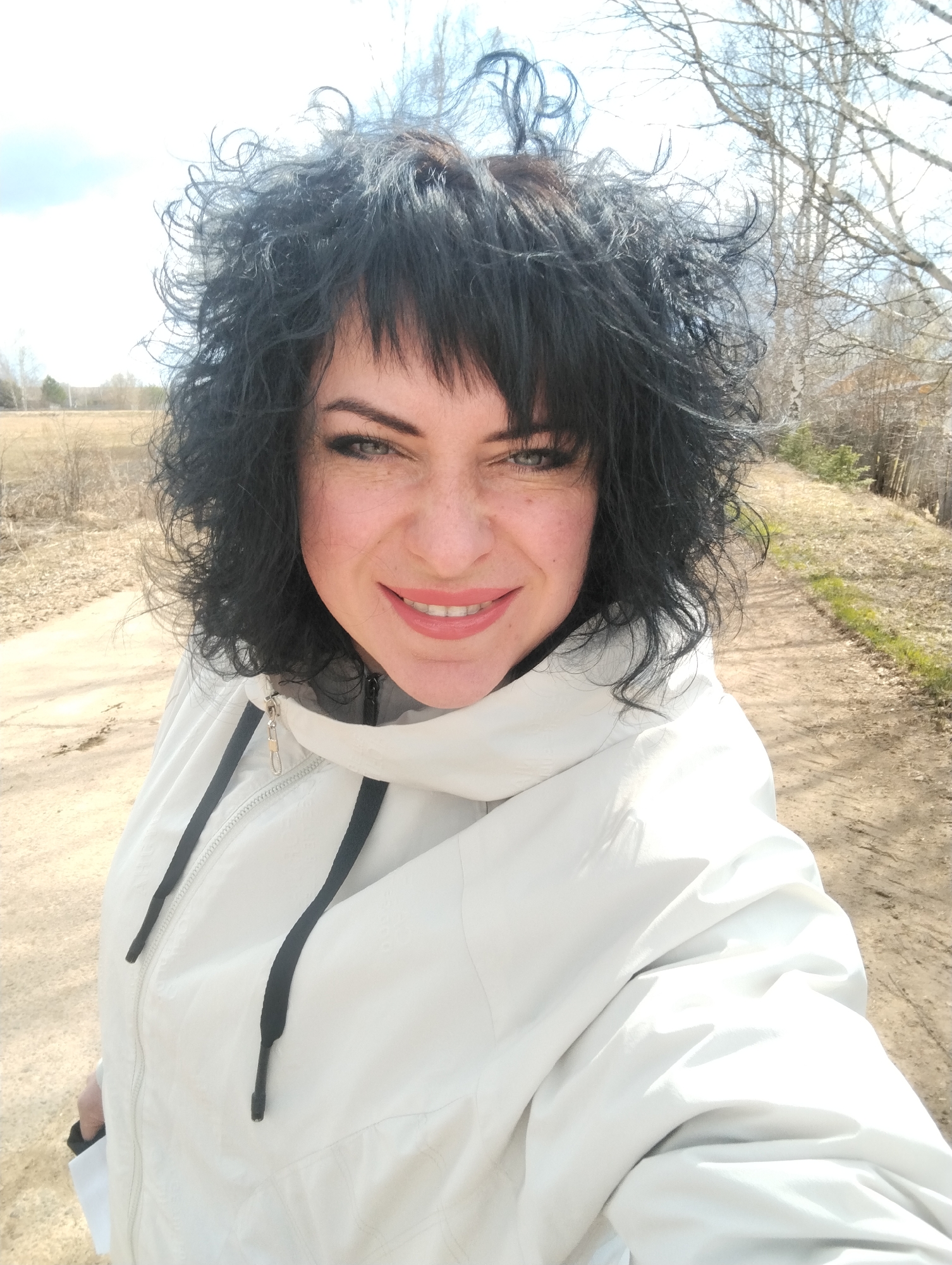 A selfie features a smiling woman outdoors in Grishanki, Russia. She has voluminous, dark, curly hair with bangs and light-colored eyes, possibly blue or green, accented with dark eyeliner and mascara. Visible freckles dot her cheeks, and her lips are painted with a bright pinkish-coral lipstick as she smiles directly at the camera, revealing her teeth.
She wears a light-colored, likely white or off-white, hooded jacket. The jacket has a silver zipper and black drawstrings around the neck and hood. The lighting is bright and natural, suggesting a sunny day, with her hair appearing slightly wind-swept.
The background reveals a rural or semi-rural landscape. To the woman's right, a dirt path stretches into the distance, bordered by dry, sparse vegetation and bare deciduous trees, characteristic of an early spring setting. The sky above is bright and partly cloudy. The overall mood is cheerful and open-air. No visible text is present in the image.
