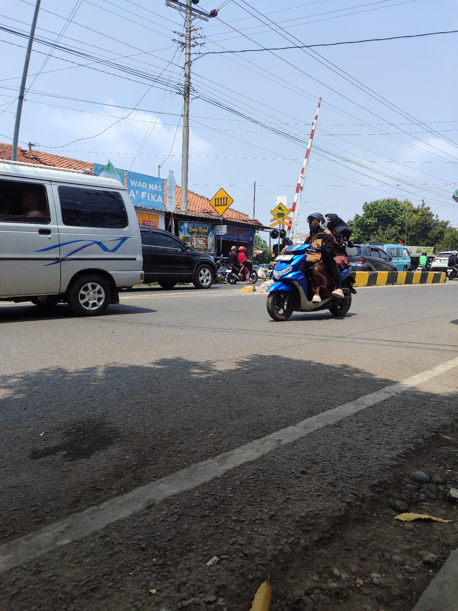 This is a street-level, eye-level, full-shot photograph taken on a sunny day. The scene depicts a busy road in Tegal, Indonesia, with a railway crossing in the background. A blue scooter with two passengers, both women wearing hijabs, is the primary focus in the foreground, heading towards the right of the frame. The driver wears a helmet and a black mask, while the passenger is dressed in a brown hijab and dress.
To the left of the scooter, a white van is parked. Behind the van, a black SUV is visible. Further in the background, a blue van is seen. A railway crossing gate, painted with red and white stripes, is raised, indicating that a train is not currently passing. Several traffic signs are present, including a yellow diamond-shaped sign with a train symbol and another with a down arrow.
The road surface is asphalt, with a white line marking the lane. The environment includes a sidewalk on the right side of the road, with some vegetation and rocks. Buildings with tiled roofs line the street in the background. The sky is clear and blue with some power lines crisscrossing overhead, characteristic of urban infrastructure in Indonesia. The overall impression is one of a typical, active street scene in Tegal.
