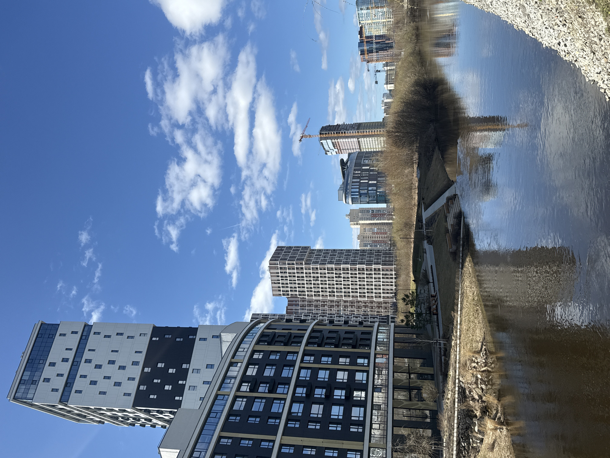 The image depicts a daytime scene in Yekaterinburg, Russia, featuring modern architecture along a riverfront. A building with a cantilevered section and geometric pattern stands prominently on the left, with neighboring buildings of varying heights and designs extending into the background. A construction crane is visible, indicating ongoing development. The sky is blue with scattered white clouds. To the right, a river reflects the sky and the buildings. The riverbank is lined with trees and a stone embankment.