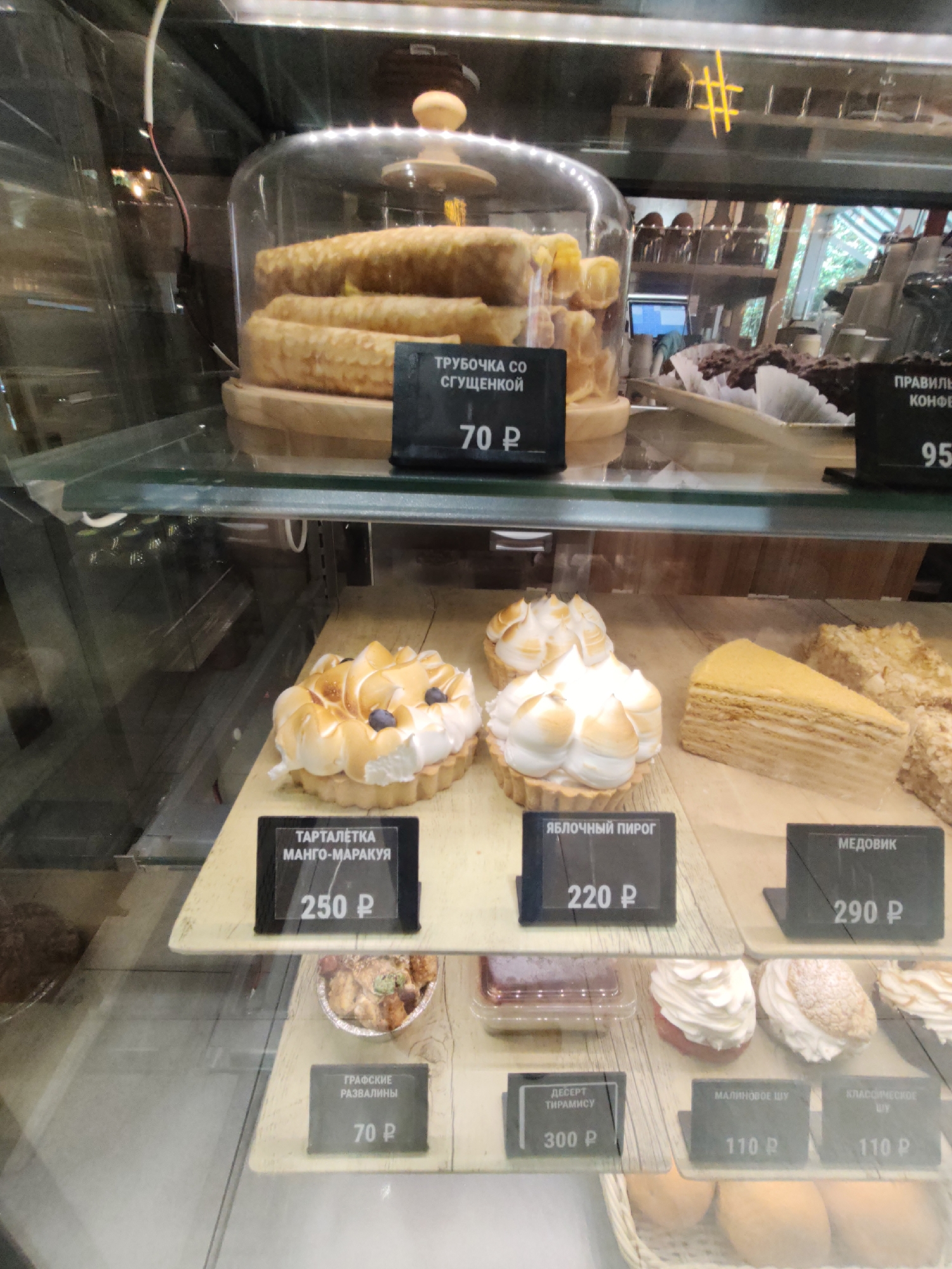 The image shows a display case in a bakery or cafe. The display case contains various desserts, including pastries, cakes, and possibly some savory items. The items are arranged on shelves and on wooden boards. Many of the desserts have small black signs in front of them with prices and names written in Cyrillic.
In the top shelf, there are several cylindrical pastries that appear to be crepes filled with cream or some other filling. A sign in front of them reads "ТРУБОЧКА СО СГУЩЕНКОЙ" which translates to "Tube with condensed milk" and the price is "70 P". To the right, there is a sign that is partially visible, but the word "ПРАВИЛЬНЫЙ" (Proper) is visible, possibly indicating a type of confectionery. The price is "95 P".
The middle shelf displays more elaborate desserts. On the left, there is a tartlet with meringue and what appears to be blueberries. The sign reads "ТАРТАЛЕТКА МАНГО-МАРАКУЙЯ" (Mango-Passion Fruit Tartlet) with a price of "250 P". Next to it, there is a round pastry with meringue, and a sign for "ЯБЛОЧНЫЙ ПИРОГ" (Apple Pie) with a price of "220 P". To the right of that, a slice of layered cake, likely a honey cake ("МЕДОВИК" - Medovik), is displayed with a price of "290 P". Further to the right, another cake slice is visible.
The bottom shelf shows smaller desserts. On the left, there is a sign for "ГРАФСКИЕ РАЗВАЛИНЫ" (Grafskie Razvaliny, a type of cake) with a price of "70 P". In the center, a dessert labeled "ДЕСЕРТ ТИРАМИСУ" (Dessert Tiramisu) is priced at "300 P". To the right of the tiramisu, there are two pastries, possibly cream puffs or eclairs, with signs indicating "МАЛИНОВОЕ ШУ" (Raspberry Shu) and "КЛАССИЧЕСКОЕ ШУ" (Classic Shu), both priced at "110 P".
The overall environment appears to be indoors, with artificial lighting. The background shows more shelves with products and possibly seating areas. There are no people clearly visible in the frame. The image is taken from a slightly elevated angle looking down into the display case.
Based on the visual cues (bakery display case with Cyrillic signage), this image is likely from a cafe or bakery in Russia. The specific location in Sochi, Russia cannot be determined from the image alone.