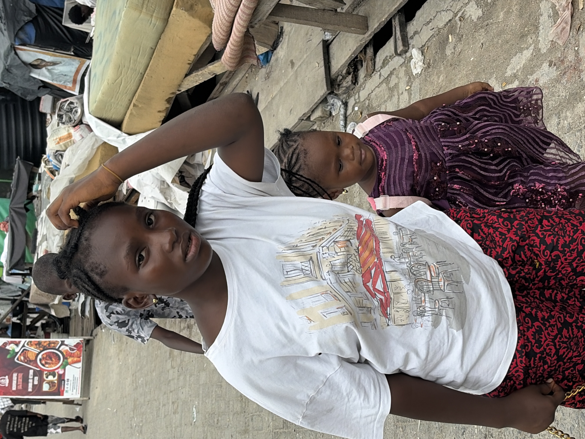 A young Black girl, with braided hair and wearing a white t-shirt with a graphic print and red patterned pants, stands in the foreground of the image. She is touching her head with her right hand, which is adorned with a yellow bracelet. To her right and slightly behind her, a younger Black girl in a shimmering purple dress with pink straps is looking down. In the background, other children and adults are visible, along with market stalls, debris, and a framed picture. The scene appears to be outdoors in a bustling urban environment, consistent with the city of Igboefon, Nigeria.
