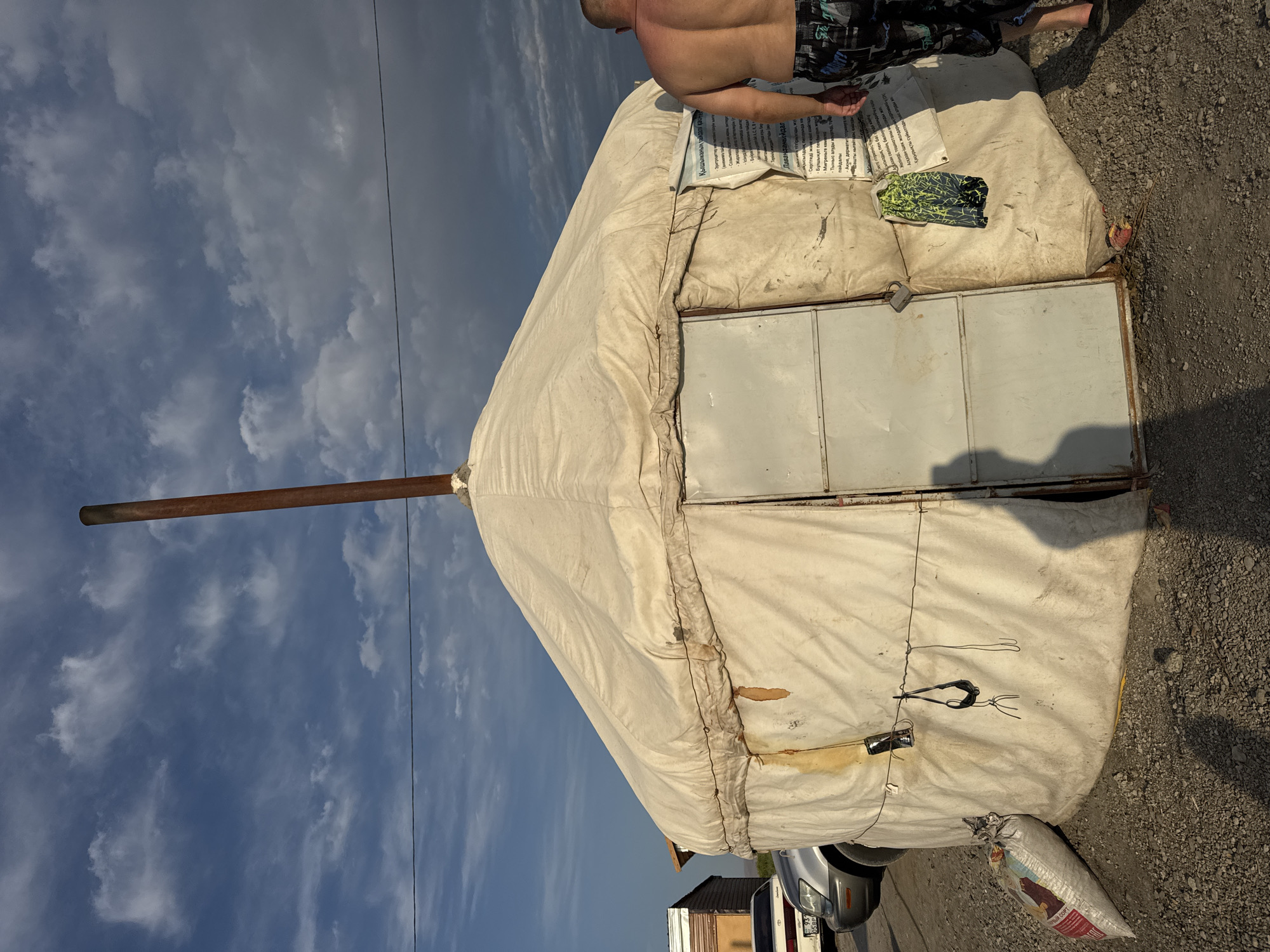 The image depicts a yurt, a traditional nomadic dwelling, set in what appears to be an outdoor or semi-rural environment. The yurt is constructed from a light-colored fabric, likely canvas, stretched over a circular frame. A metal chimney pipe protrudes from the apex of the yurt's conical roof. A metal door is visible on the front of the yurt, appearing somewhat weathered.
To the left of the yurt, a silver car is parked, with part of its front visible. Behind the car, a small, light-colored building with horizontal siding can be seen. A white sack, possibly containing grain or feed, rests on the ground near the car and the yurt. A thin wire runs horizontally across the scene, secured to the yurt and likely to other support structures.
On the right side of the image, a man is partially visible, walking away from the yurt. He is shirtless, revealing his back, and is wearing patterned shorts. His feet are visible, and he appears to be walking on a gravel or dirt surface.
A sign, appearing to be made of paper or thin plastic, is affixed to the side of the yurt near the door. The text on the sign is in a foreign language and is difficult to read clearly in the provided image. The sky above is partly cloudy, with a mix of blue sky and darker, heavier clouds, suggesting dynamic weather conditions. The overall impression is a somewhat utilitarian setting, possibly a temporary settlement or a roadside stop.