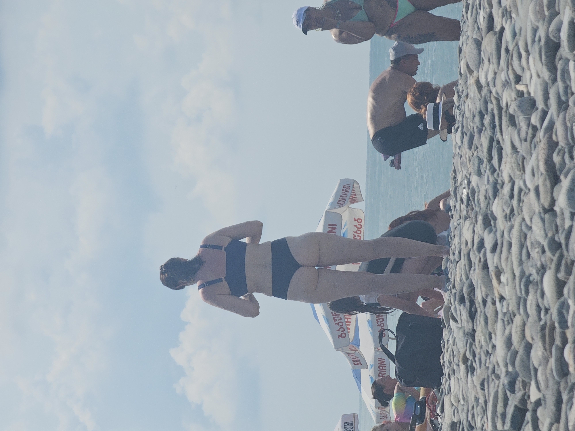 The image depicts a crowded pebble beach in Batumi, Georgia, under a hazy sky with scattered clouds. In the foreground, a woman in a dark blue bikini stands facing away from the camera, her back to the viewer. Behind her, several beach umbrellas are visible, some with Georgian script. Other beachgoers are scattered throughout the scene, with some sitting or lying on the pebbles and others in the water.
To the left, a group of people are gathered near a black duffel bag, with one woman in a brightly colored swimsuit looking to her left. In the center, a man in black shorts is bent over, appearing to interact with someone below him. To the right, a woman wearing a white cap and a light green bikini stands near the water, her hand raised to her mouth. The sea is visible in the background, with gentle waves lapping at the shore. The overall impression is one of a busy summer day at the beach.