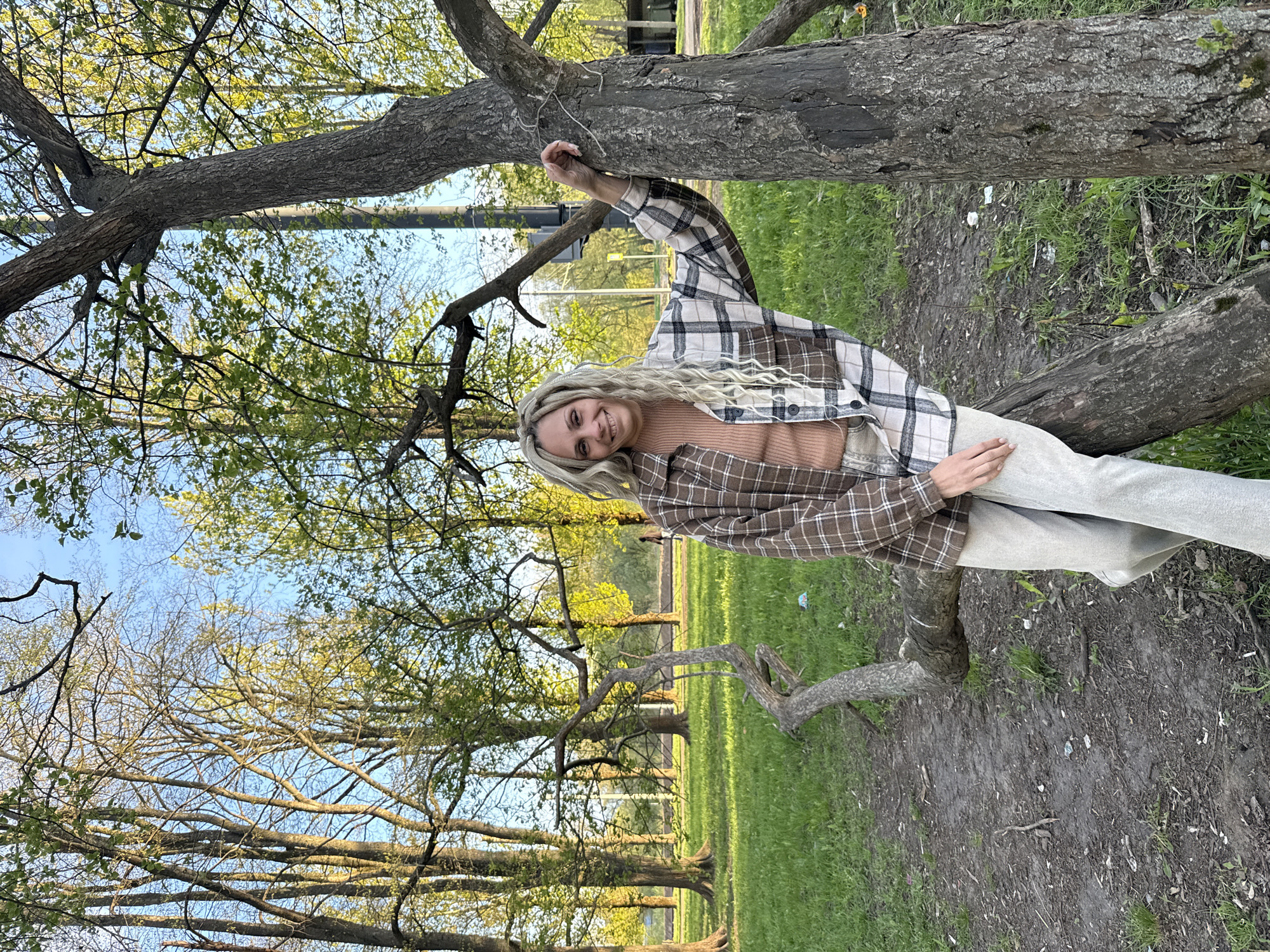A woman stands outdoors in Rusanovo, Russia, leaning against a fallen tree. She is wearing a brown and white plaid shirt over a neutral-toned top and light-colored pants. The surrounding environment consists of trees, grass, and bare earth. The sky is visible through the branches of the trees.