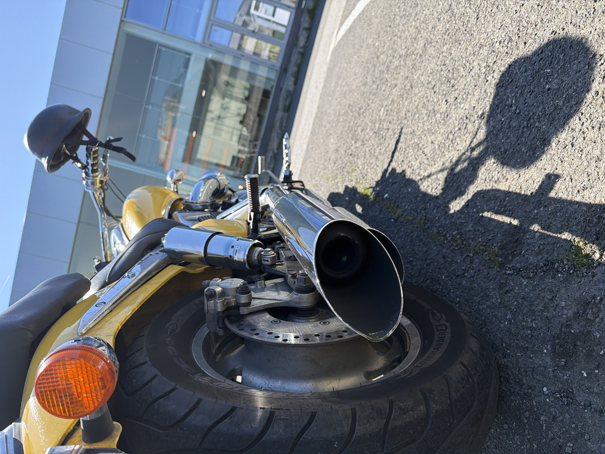 The image shows a close-up view of a yellow motorcycle parked on asphalt in an urban environment, likely in Unknown, Unknown. The motorcycle is detailed from the rear wheel to the handlebars, including the brake caliper, exhaust pipes, a rear tire, a turn signal and the seat. A helmet is mounted on the handlebars. A shadow is cast on the asphalt. In the background, a glass building is partially visible.