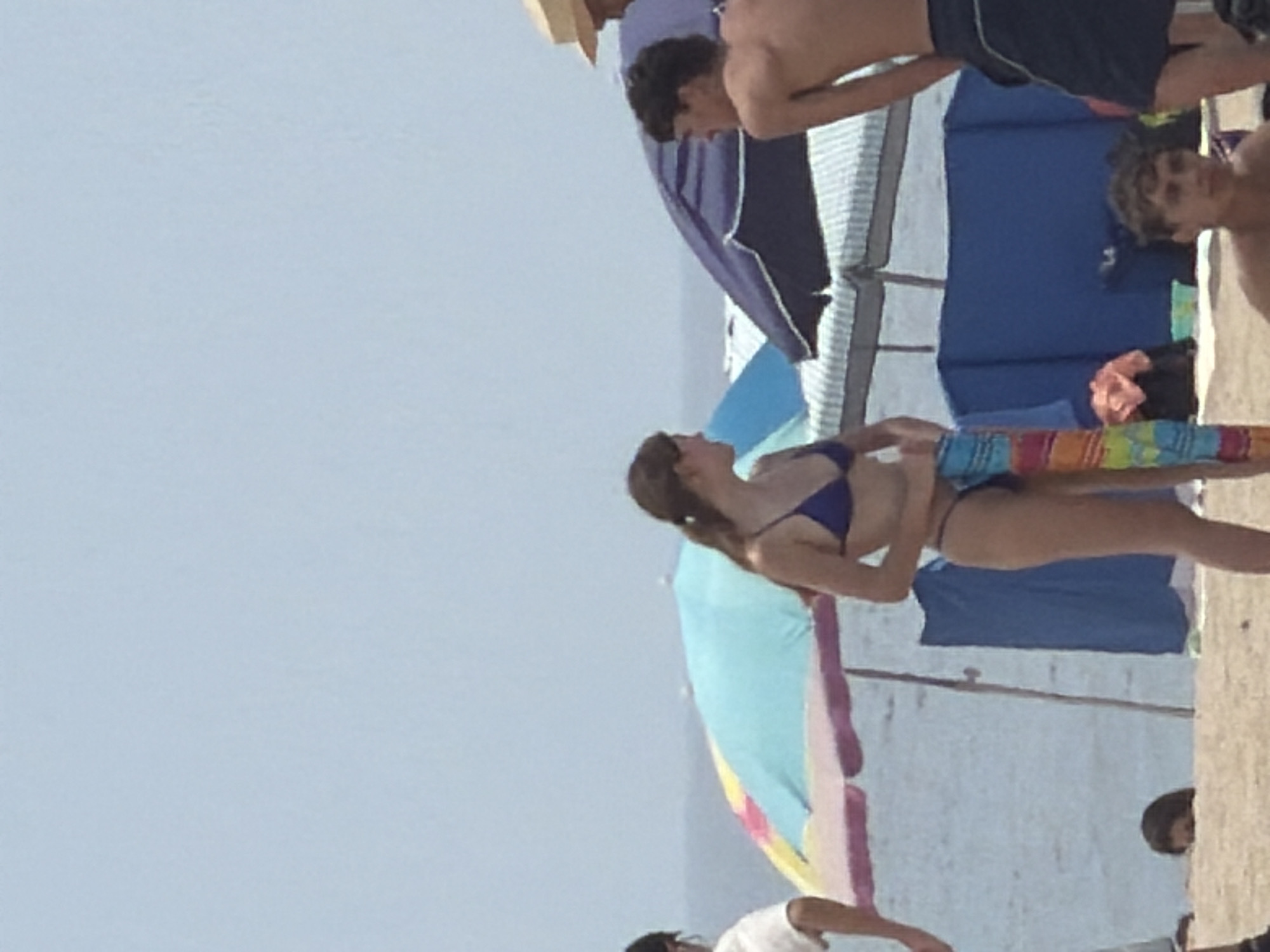 The image depicts a sunny day at the beach in Espinho, Portugal. In the foreground, a young woman in a blue bikini stands holding a colorful striped towel. To her right, a young man in dark swim trunks is partially visible from the side, his back to the camera. In the background, several beach umbrellas are set up, with one large umbrella displaying a mix of blue, yellow, and pink hues. Another dark blue umbrella is prominent behind the young man. The sandy beach stretches towards the ocean, where the water and horizon are visible under a hazy, light blue sky. Other beachgoers and beach structures are blurred in the distance, suggesting a busy atmosphere.