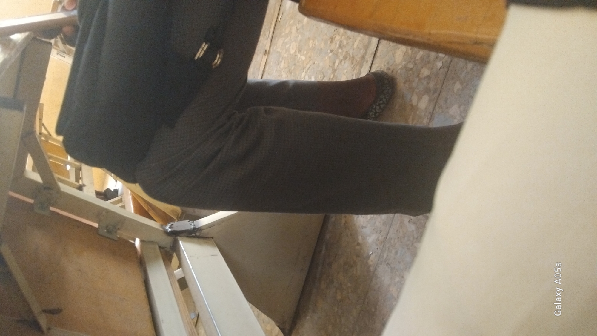 A close-up, waist-down shot captures a person seated at a table in Yali, Nigeria. The person is wearing dark grey trousers with a subtle checkered pattern and a black jacket draped over their lap. One foot is visible, clad in a dark ballet flat with a grey pattern. The background consists of the wooden surface of the table and its metal frame, a tiled floor, and the edge of another wooden table or surface. The overall lighting is dim, suggesting an indoor setting. The "Galaxy A05s" watermark is present in the lower-left corner.
