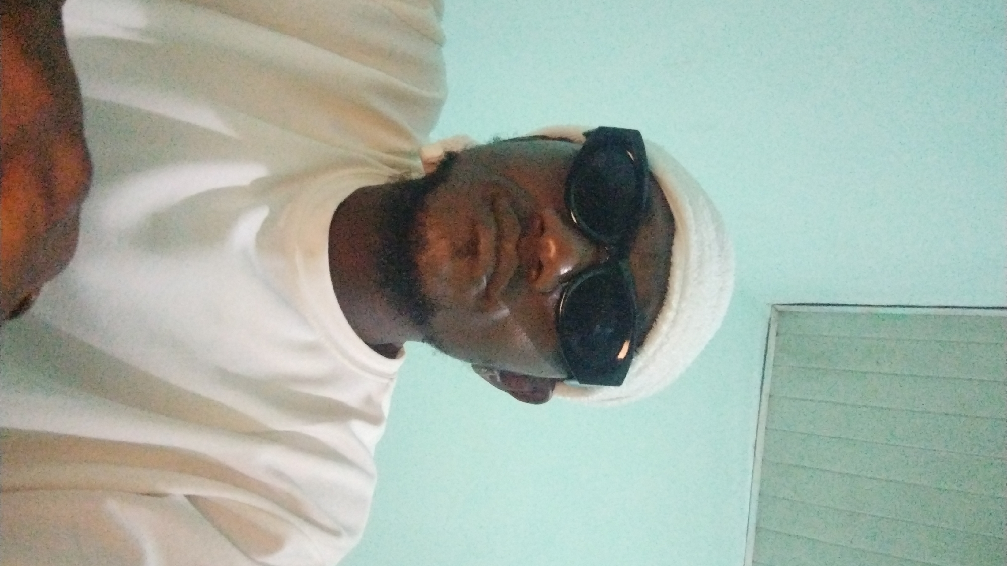 The image shows a medium shot of a person wearing a white beanie, black sunglasses, and a white t-shirt, looking directly at the camera. The background is a plain, light blue wall with a white ceiling visible in the upper right corner, which appears to be a simple suspended ceiling with horizontal panels. The lighting suggests an indoor setting. The person's facial features are partially obscured by the sunglasses. The overall impression is a casual, self-taken photograph. Without specific visual cues within the image that definitively link it to Jos, Nigeria, it is not possible to confirm its location within that city.