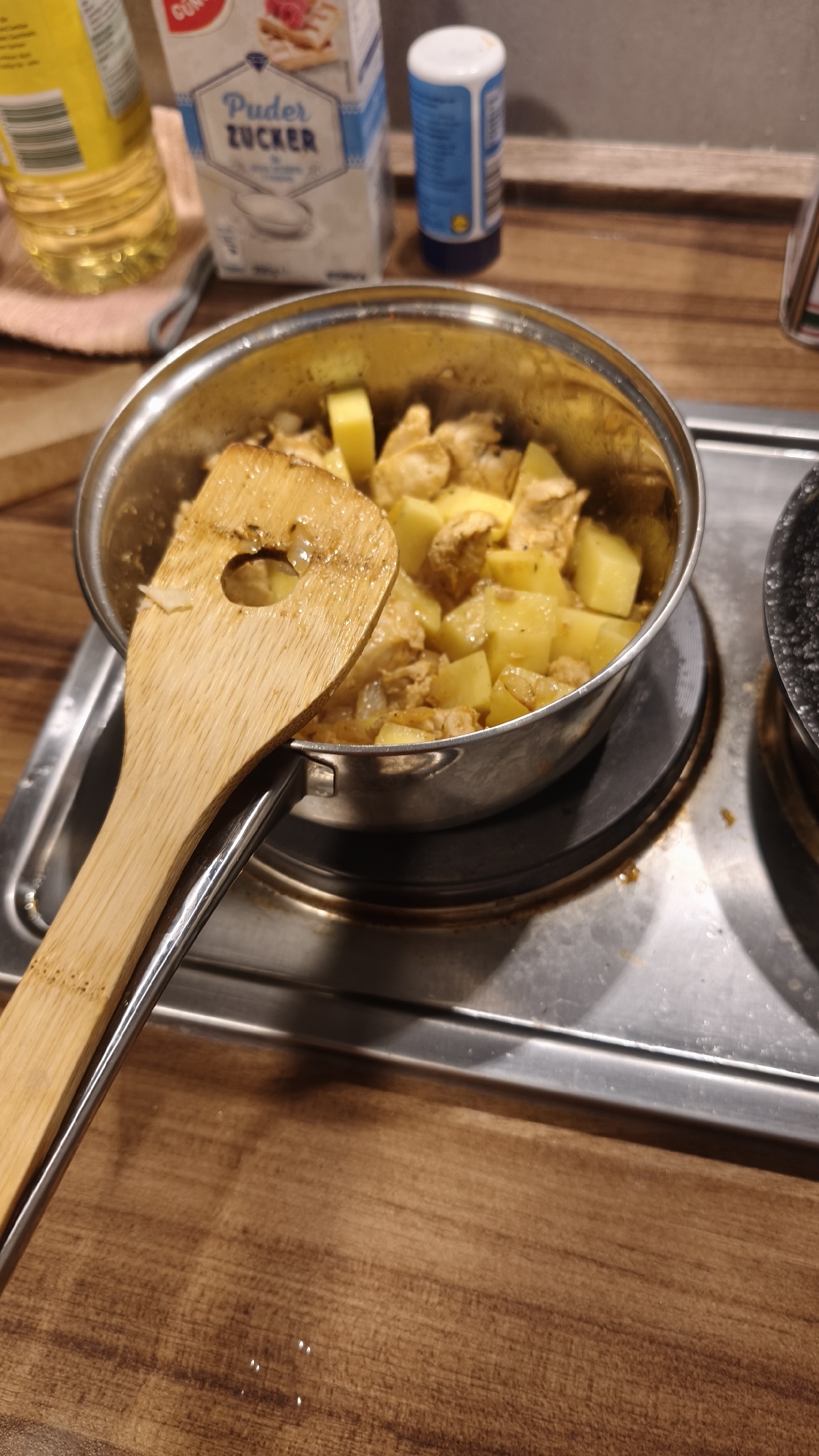 This close-up image captures a cooking scene in what appears to be a kitchen, with a focus on a metal pot on an electric stovetop. The pot contains diced yellow potatoes and pieces of light-colored meat, likely chicken, mixed in a sauce or seasoning. A wooden spatula with a hole in its center rests on the pot's handle, suggesting recent stirring or ongoing cooking activity.
In the background, several items are visible on a wooden countertop. To the left, a yellow bottle, likely cooking oil, is partially obscured. Next to it, a white package prominently displays the German text "Puder ZUCKER," indicating powdered sugar. Further to the right, a small blue and white shaker bottle is visible. A portion of another dark pot or pan can be seen on the stovetop to the right of the main pot. The electric stovetop is silver-colored, and the overall setting suggests a domestic environment where a meal is being prepared. The lighting indicates an indoor setting, but the specific time of day is not discernible. No people are visible, and no particular emotions are conveyed by the scene.