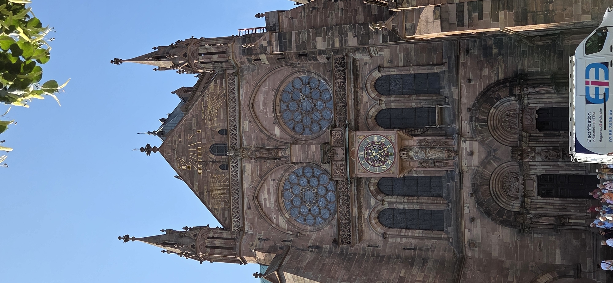 This image showcases the exterior of a grand, Gothic-style cathedral, likely in Strasbourg, France, given the context. The building is constructed from reddish-brown stone and features intricate architectural details, including pointed spires, ornate carvings, and rose windows. A prominent clock face with Roman numerals and astrological symbols is visible on the facade. A sundial is also present on the upper part of the building.
In the foreground, to the left, a branch of a tree with green leaves and small white flowers hangs into the frame, suggesting it's springtime or summer. To the right, a white van with blue lettering is parked. The text on the van reads: "EIE Electrification Industrielle de l'Est Haguenau & Molsheim" followed by a phone number "03 88 73 56 56". A group of people, appearing to be tourists, are gathered in front of the cathedral's entrance, looking up at the architecture. The sky is clear and bright blue, indicating a sunny day. The overall impression is of a bright, clear day at a significant historical and architectural landmark.