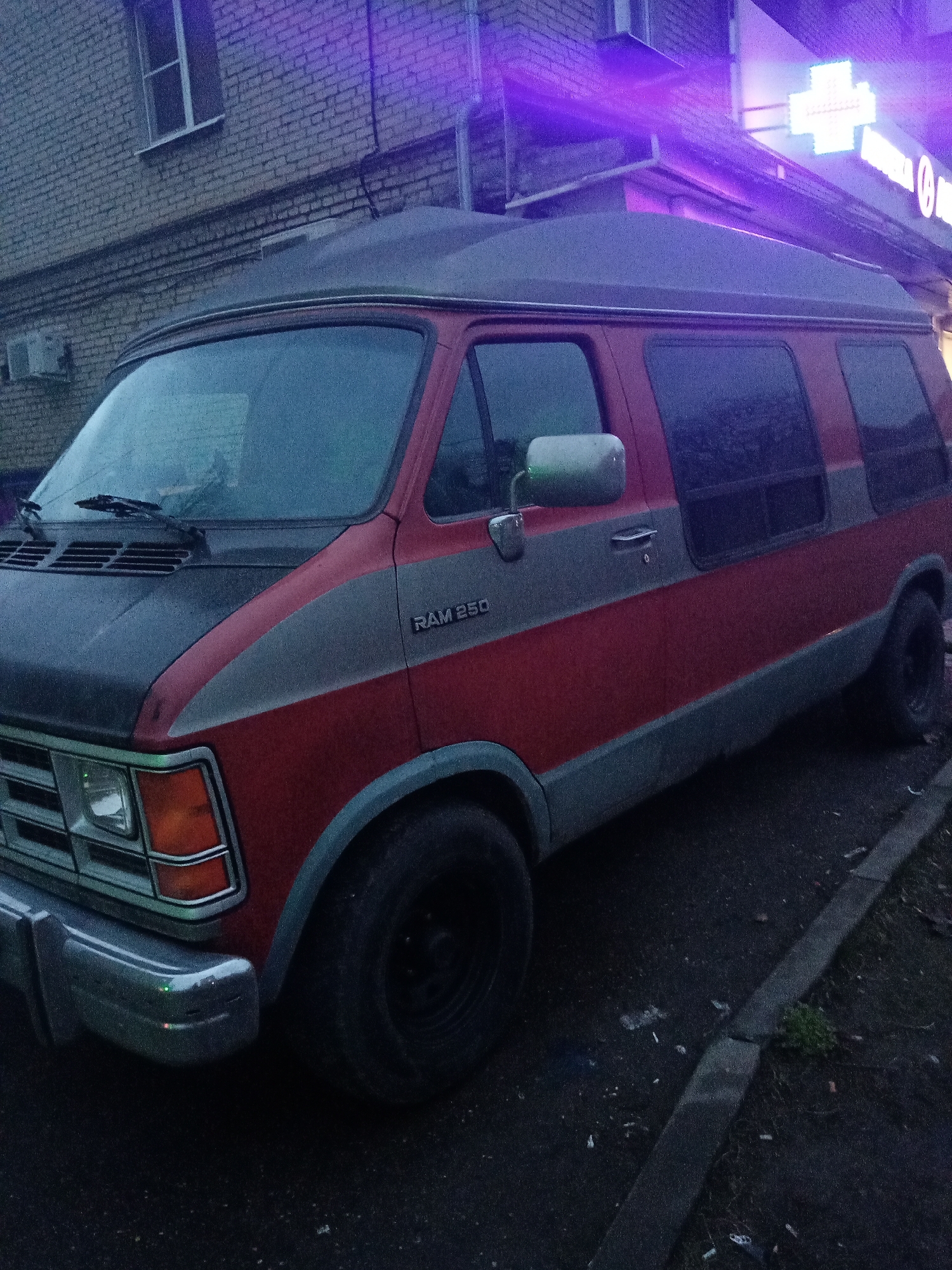 The image depicts a red and grey Dodge Ram 250 van parked on a street next to a curb. The building in the background appears to be brick, with a lit medical cross sign visible on a building facade. The lighting suggests it is evening or dusk. Based on the presence of Russian text (possibly "Elektrostal") and the architectural style, the location is likely within or near the city of Elektrostal, Russia. No people are visible in the image. The van is the primary subject. The environment includes the street, curb, and surrounding buildings.