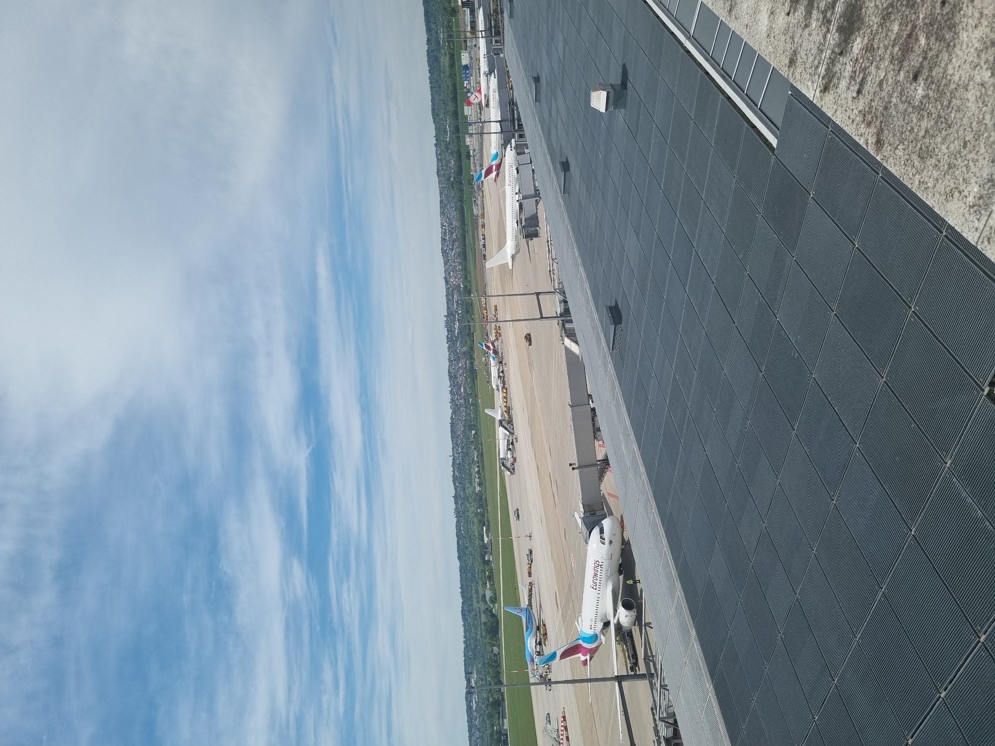 The image shows an airport scene in Echterdingen, Germany. Several airplanes are visible on the tarmac, including a Eurowings aircraft. The sky is partly cloudy. A building with a dark grey, patterned exterior wall is in the foreground.