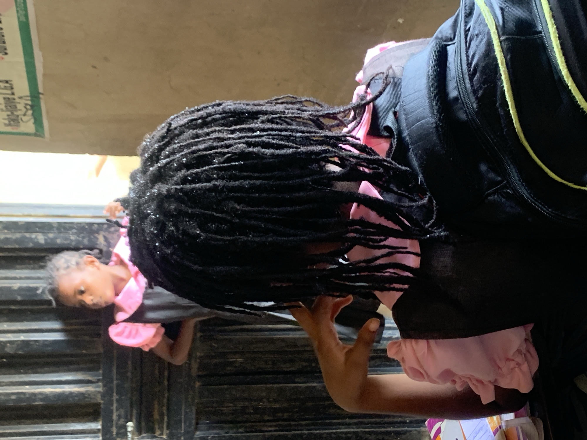 A young girl with dreadlocks stands near an open metal gate. She wears a school uniform consisting of a dark gray dress with pink trim, carrying a black backpack with a yellow stripe. Her younger sister, also in a school uniform, looks up at her from behind the gate. The scene appears to be in the city of Unknown, Unknown.