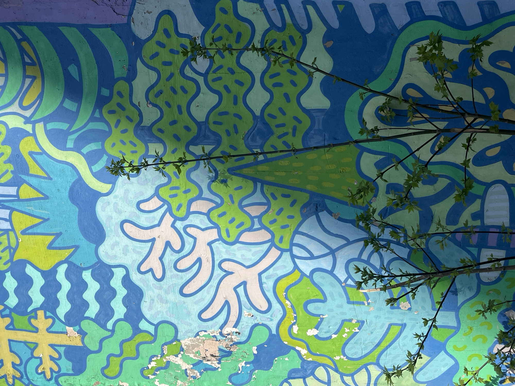 The image depicts a colorful mural on a wall, partially obscured by tree branches. The mural features abstract shapes and patterns in shades of blue, green, yellow, and white. The paint is peeling in some areas, revealing the underlying wall surface. Tree branches with small green leaves are visible in the foreground, partially covering the mural. The location is Unknown, Unknown.