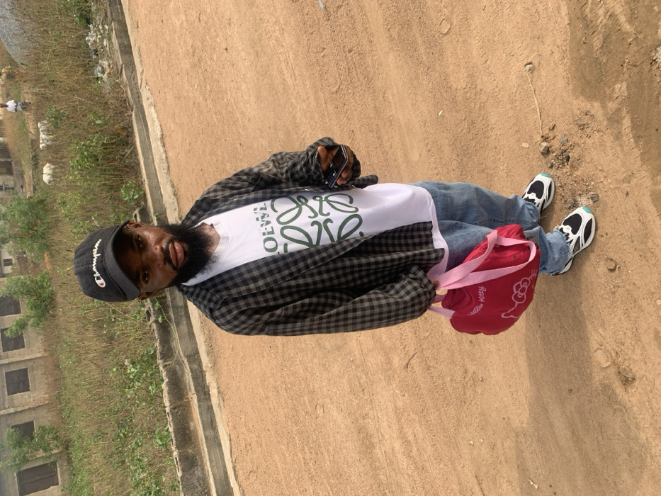 A male individual is observed standing on an unpaved, reddish-brown dirt road in Eika-Adagu New Layout, Nigeria. He presents with characteristics of dwarfism. The individual wears a black "Champion" baseball cap, a white t-shirt featuring a green "LOEWE" logo, an open dark-grey and white checkered long-sleeve shirt, light-blue jeans, and black, white, and light-blue sneakers. In his left hand, he holds a black smartphone. In his right hand, he carries a bright pink "Hello Kitty" themed bag with pink straps. His gaze is directed towards the camera, maintaining a neutral expression. To the immediate left, a low concrete curb borders an elevated area covered with dry, tall grass and weeds. In the background, several unfinished or dilapidated concrete block structures with dark openings are visible amidst the dry vegetation. Further in the distance, two indistinct figures, one human and one animal (possibly a goat or sheep), are discernible against the landscape. The scene is illuminated by natural daylight.