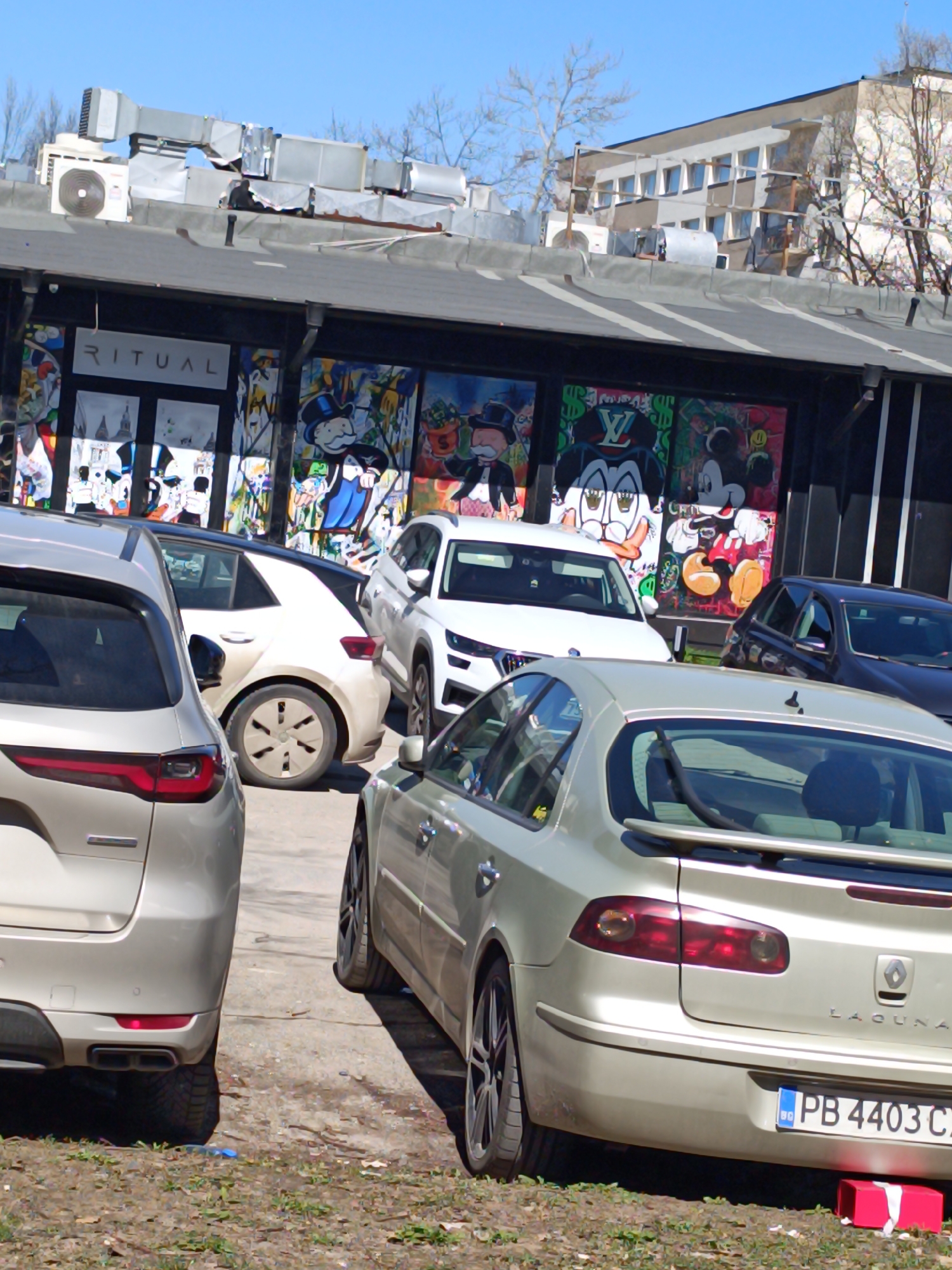 The image shows a street scene in Sofia, Bulgaria. Several cars are parked in front of a building with large, colorful murals depicting Monopoly and Mickey Mouse characters. The building has a sign reading "RITUAL". The sky is blue and clear. The car closest to the viewer is a silver Renault Laguna with a Bulgarian license plate reading "PB 4403 CЗ". There is a red object on the ground near the car, possibly a gift or package. The area appears to be a parking lot or public space with mixed pavement and patches of grass.