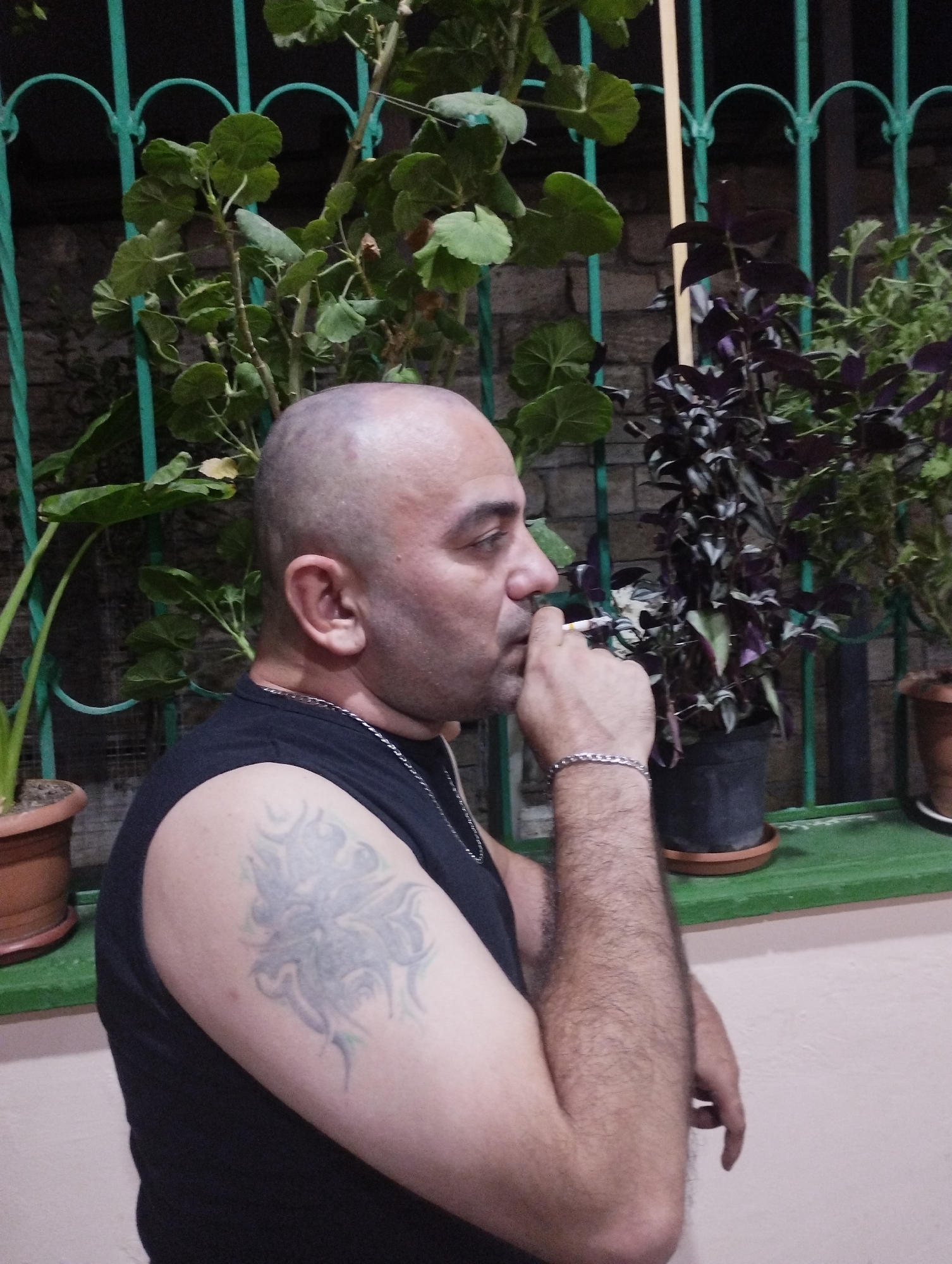 A man with a shaved head is smoking a cigarette outdoors, positioned in the foreground with his right side facing the camera. He is wearing a black sleeveless shirt and a silver chain necklace. A tattoo is visible on his left upper arm, and he wears a silver bracelet on his right wrist. He is standing in front of a green metal fence and several potted plants. The background shows a brick wall and more foliage. The image is likely taken in Bilajari, Azerbaijan.