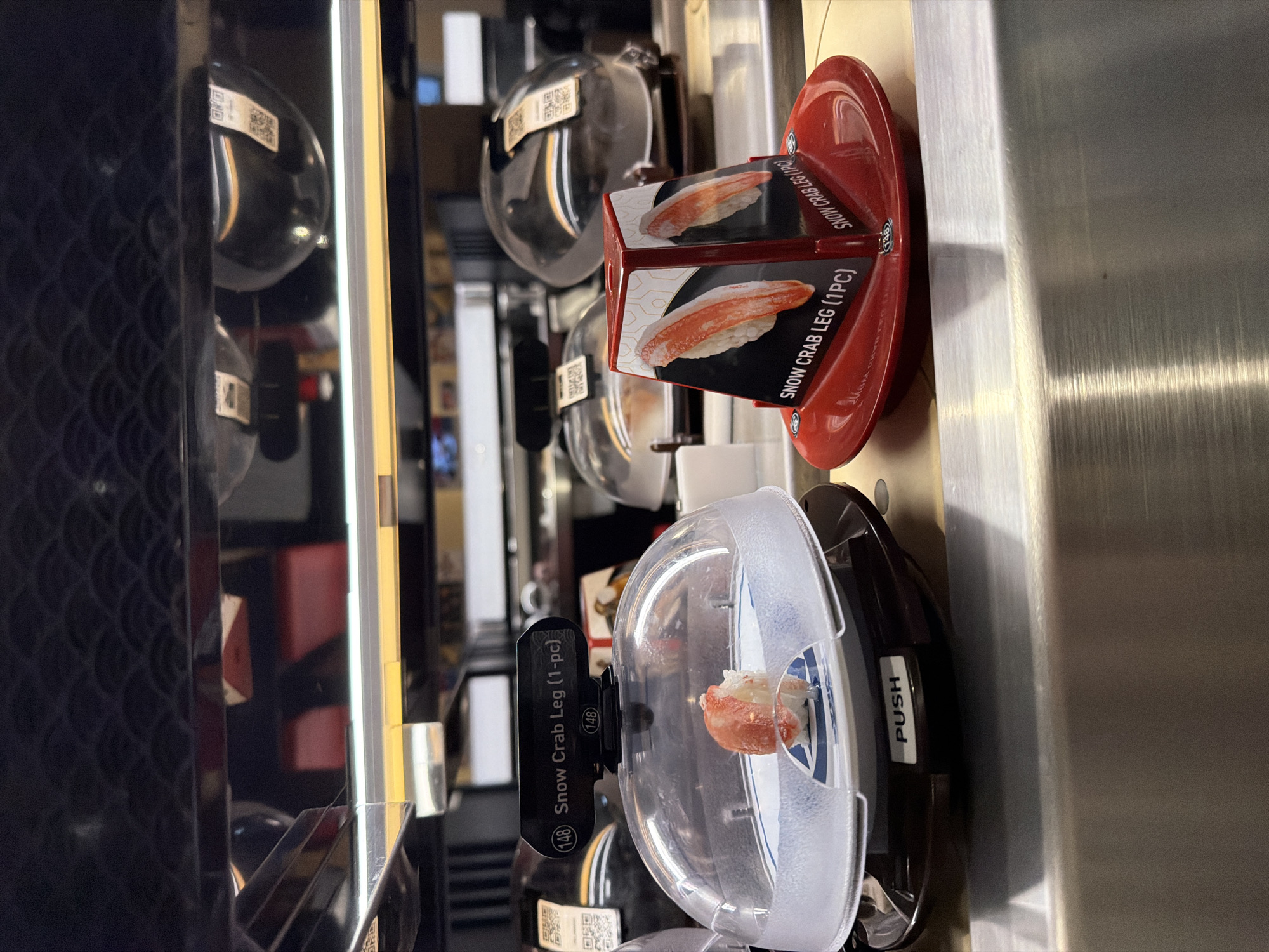The image depicts a close-up, eye-level view of a conveyor belt sushi display at a restaurant, likely in Beaverton, Oregon, United States of America. Two sushi plates are prominently featured. The plate on the left is covered with a clear dome and contains a single piece of sushi, appearing to be crab or shrimp. A black label above the dome reads "(148) Snow Crab Leg (1-pc)". The plate on the right is red and is topped with a triangular display featuring images of snow crab legs and the text "SNOW CRAB LEG (1PC)" on each visible face. The background shows blurred shelves with more covered sushi plates, suggesting a busy sushi bar environment. The lighting is artificial, illuminating the sushi and the metallic surfaces of the display.