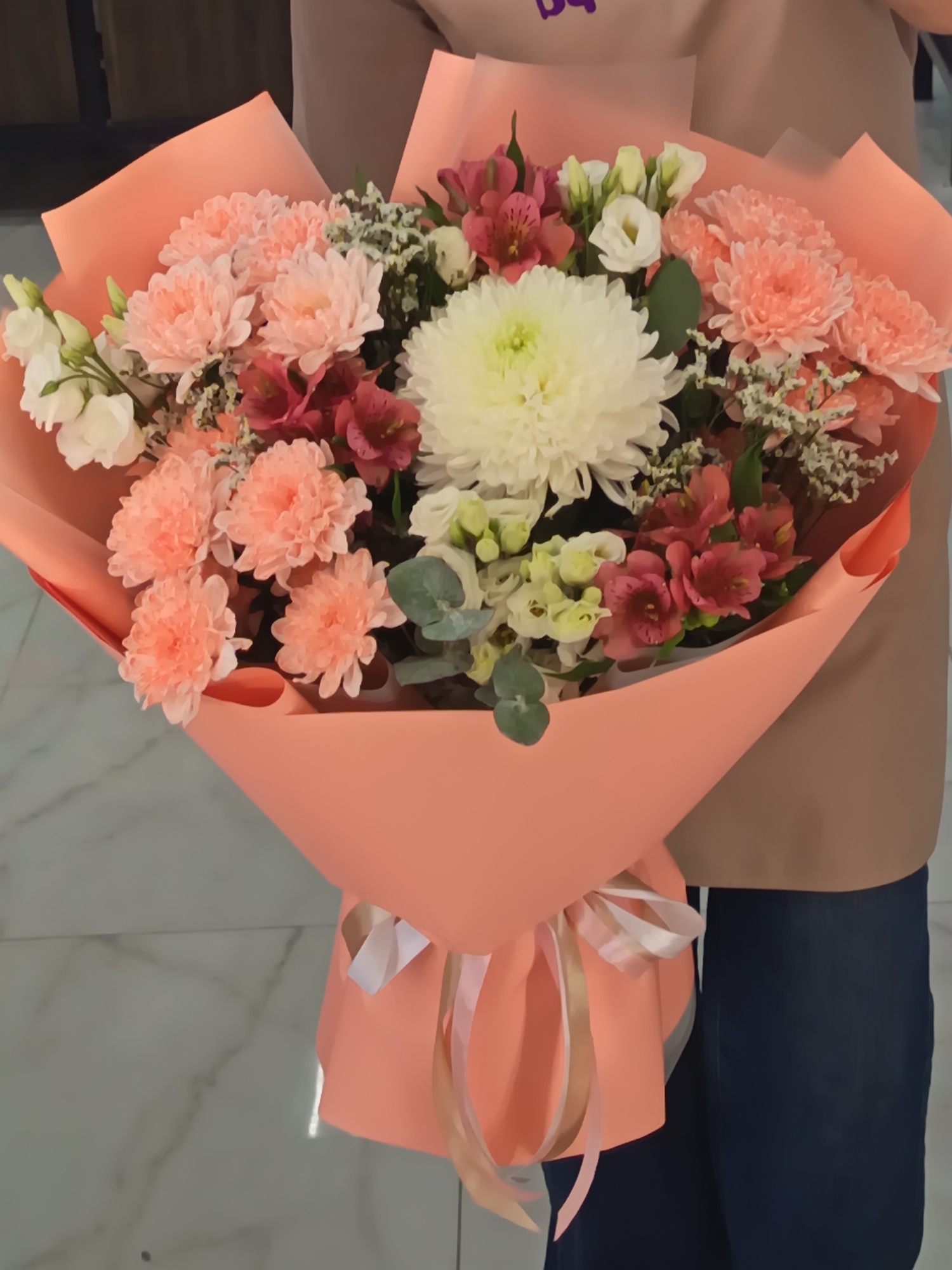 A person holds a bouquet of flowers wrapped in peach-colored paper and tied with ribbons. The bouquet contains peach and white chrysanthemums, red alstroemeria, white lisianthus, and baby's breath. The person is wearing a tan top and blue jeans, and they are standing indoors on a marble floor. The location is inferred to be Nizhnevartovsk, Russia, based on the image source.