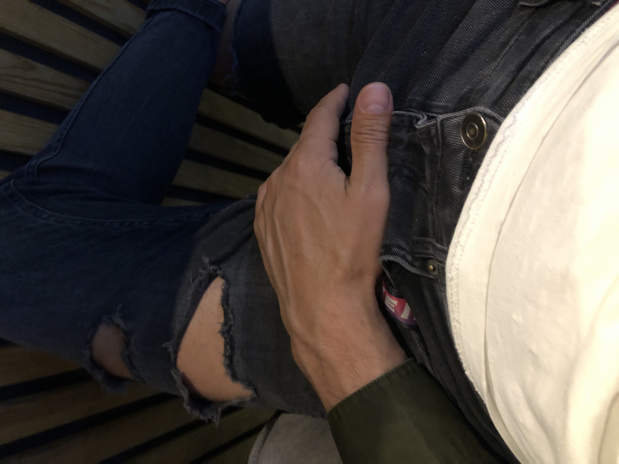 The image shows a close-up view of a person's legs and hand. The person is wearing ripped dark wash denim jeans and a white t-shirt. A hand with visible veins rests on the person's lap, over the jeans. The background consists of wooden slats, possibly from a bed frame or bench. The image does not provide any specific details that would indicate its location in Saint Petersburg, Russia.